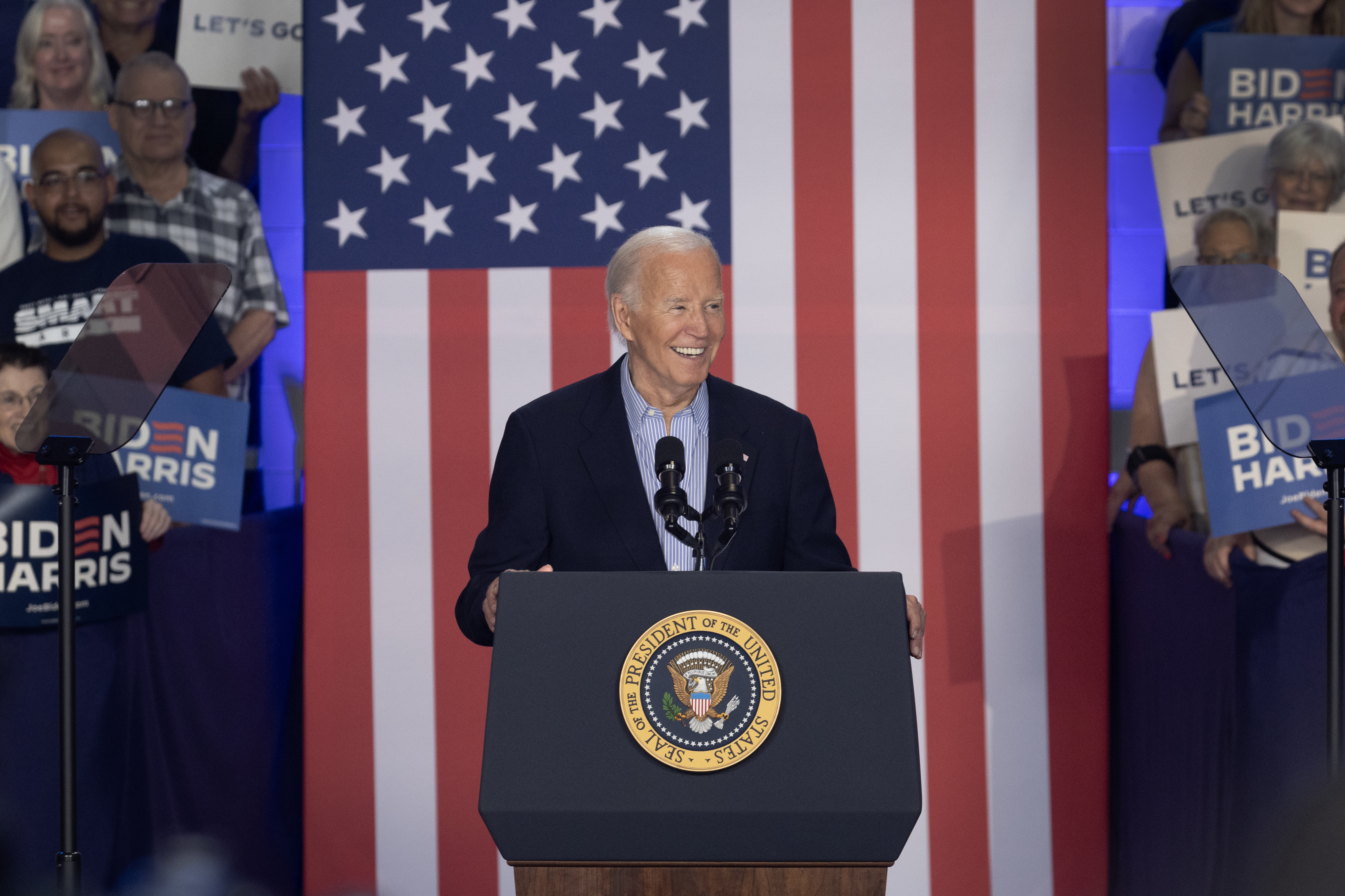 President Joe Biden speaking at a podium with an American flag backdrop. Audience members hold &quot;Biden-Harris&quot; and &quot;Let's Go&quot; signs