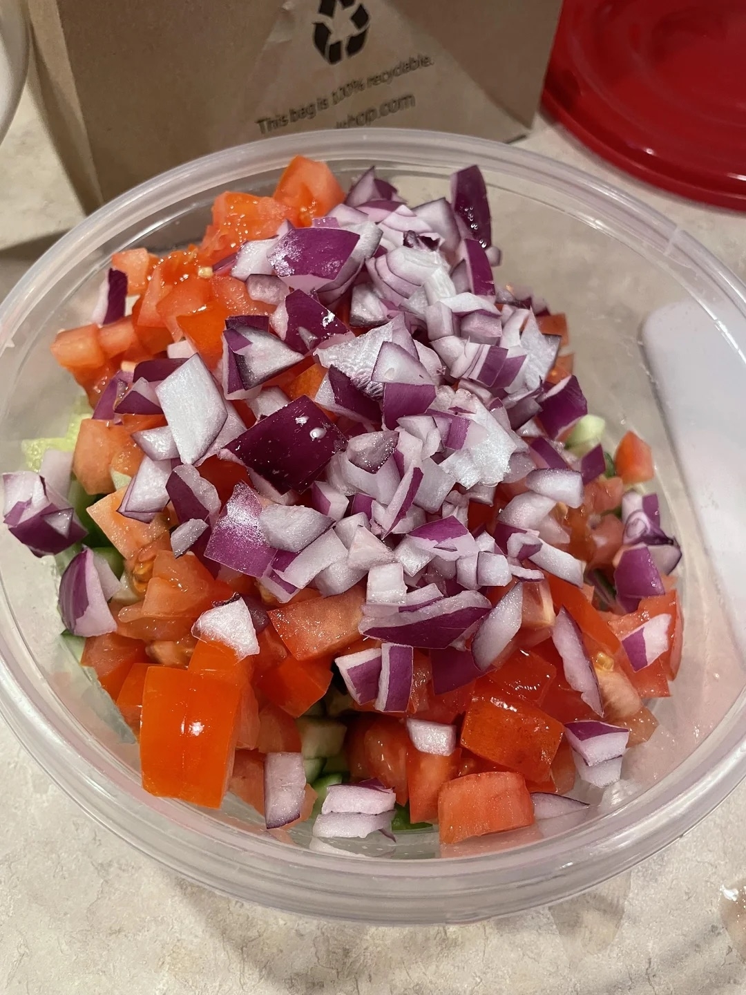 A clear plastic bowl filled with chopped red onions, tomatoes, and cucumbers on a kitchen counter
