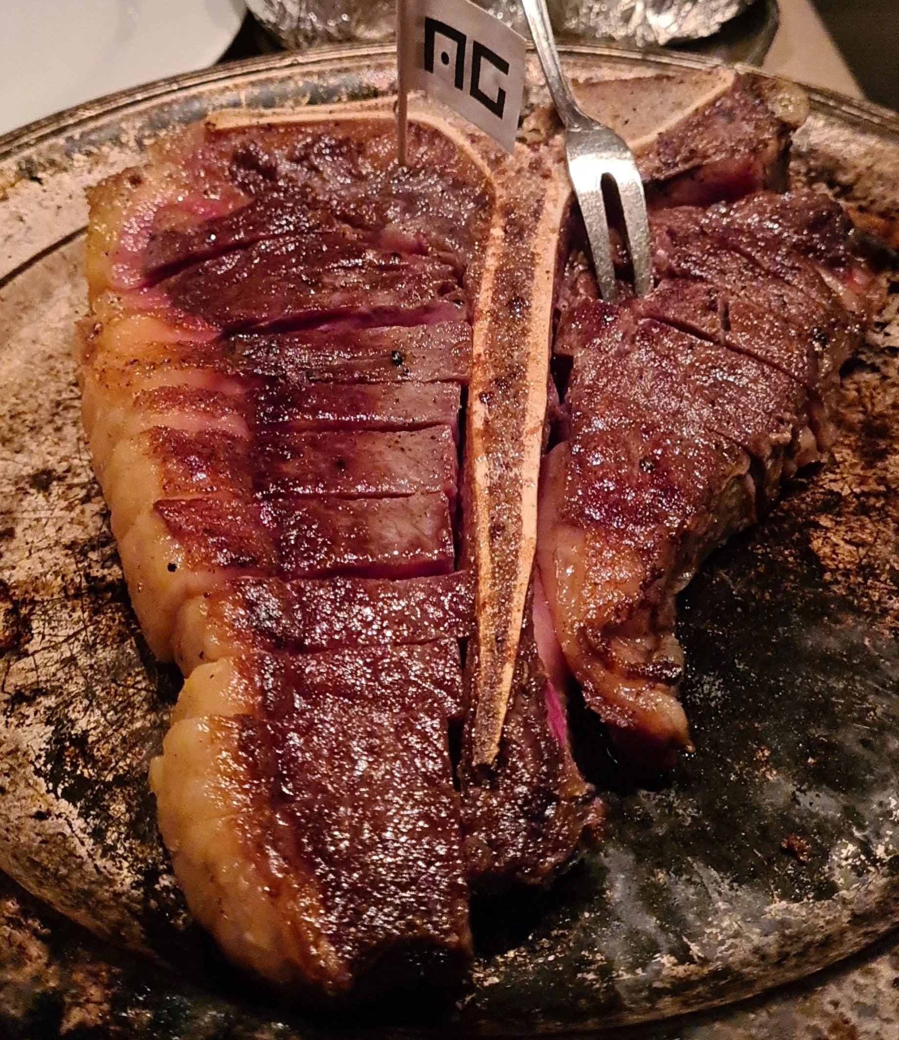 A juicy, grilled steak with a bone on a plate, partially sliced with silver utensils inserted. Two foil-wrapped baked potatoes are in the background