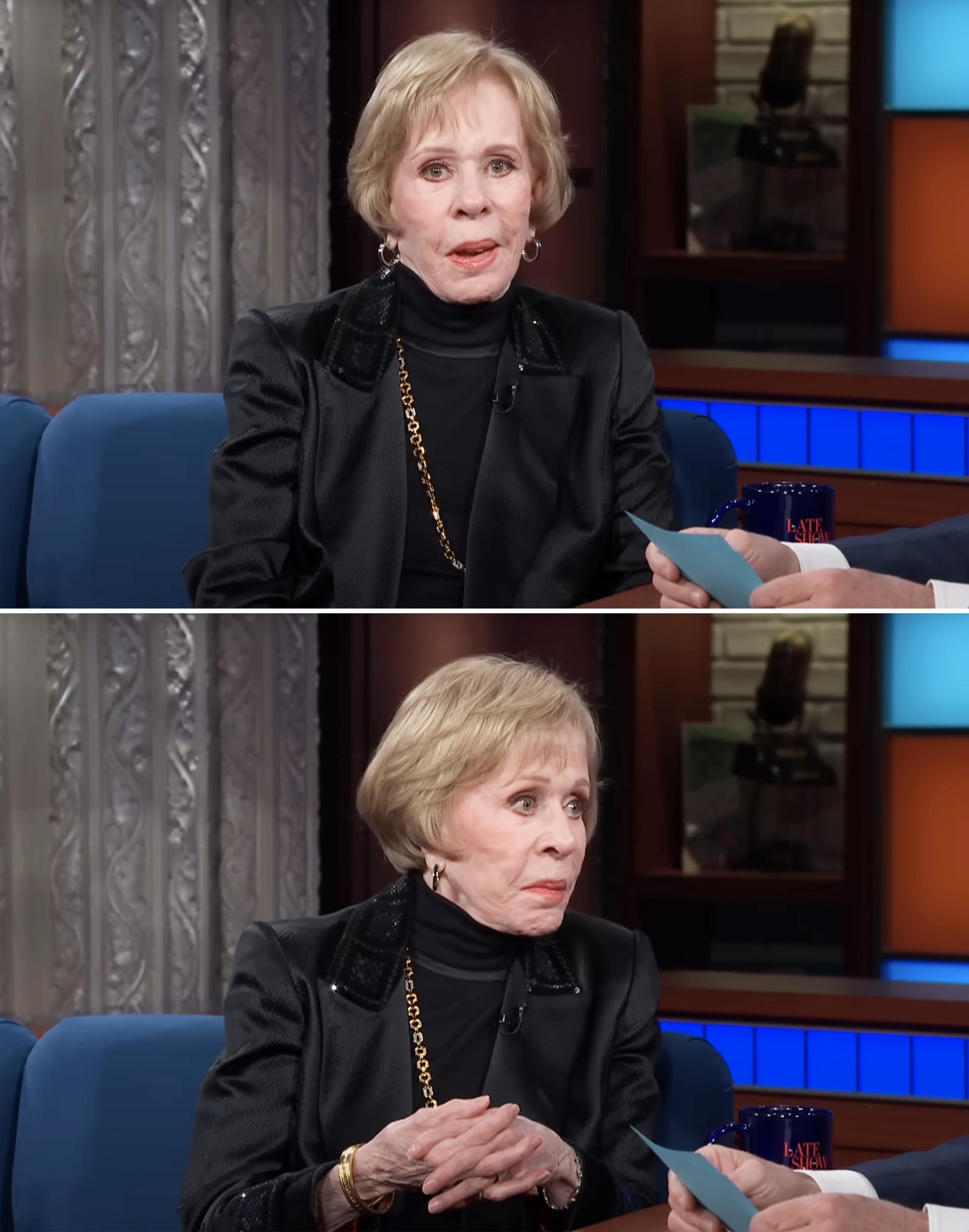 Carol Burnett, wearing a black outfit with a gold necklace, is being interviewed on a talk show. She appears engaged in conversation