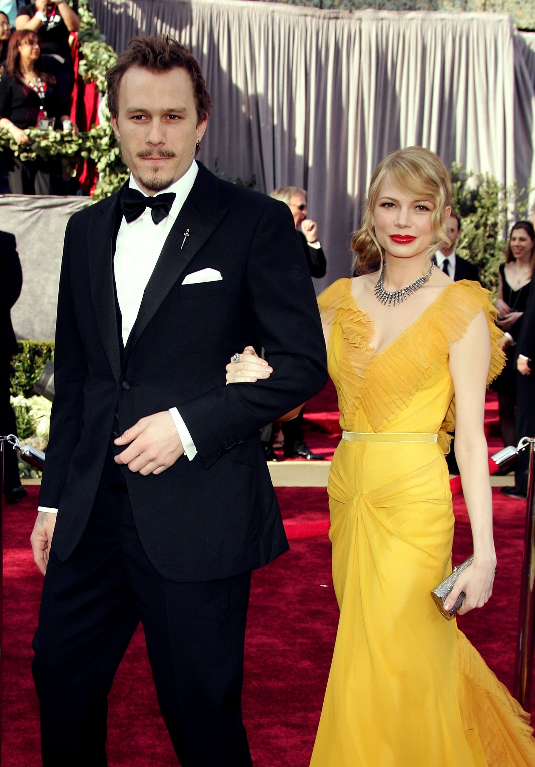 Heath Ledger and Michelle Williams on the red carpet. Heath is in a black tuxedo, and Michelle is in a yellow gown with a deep neckline and ruffled details