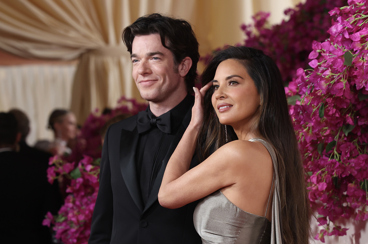 John Mulaney and Olivia Munn pose together on a red carpet event. John wears a classic black tuxedo, and Olivia wears a silver gown with a draped back