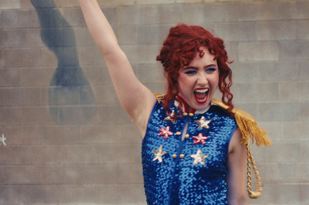 Woman with curly red hair wearing a sequined outfit with star patterns, smiling energetically and raising one arm triumphantly