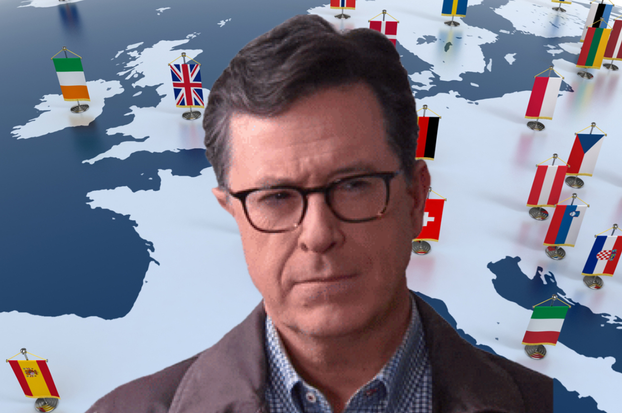 Stephen Colbert in front of a world map with various national flags, wearing a grey jacket and glasses