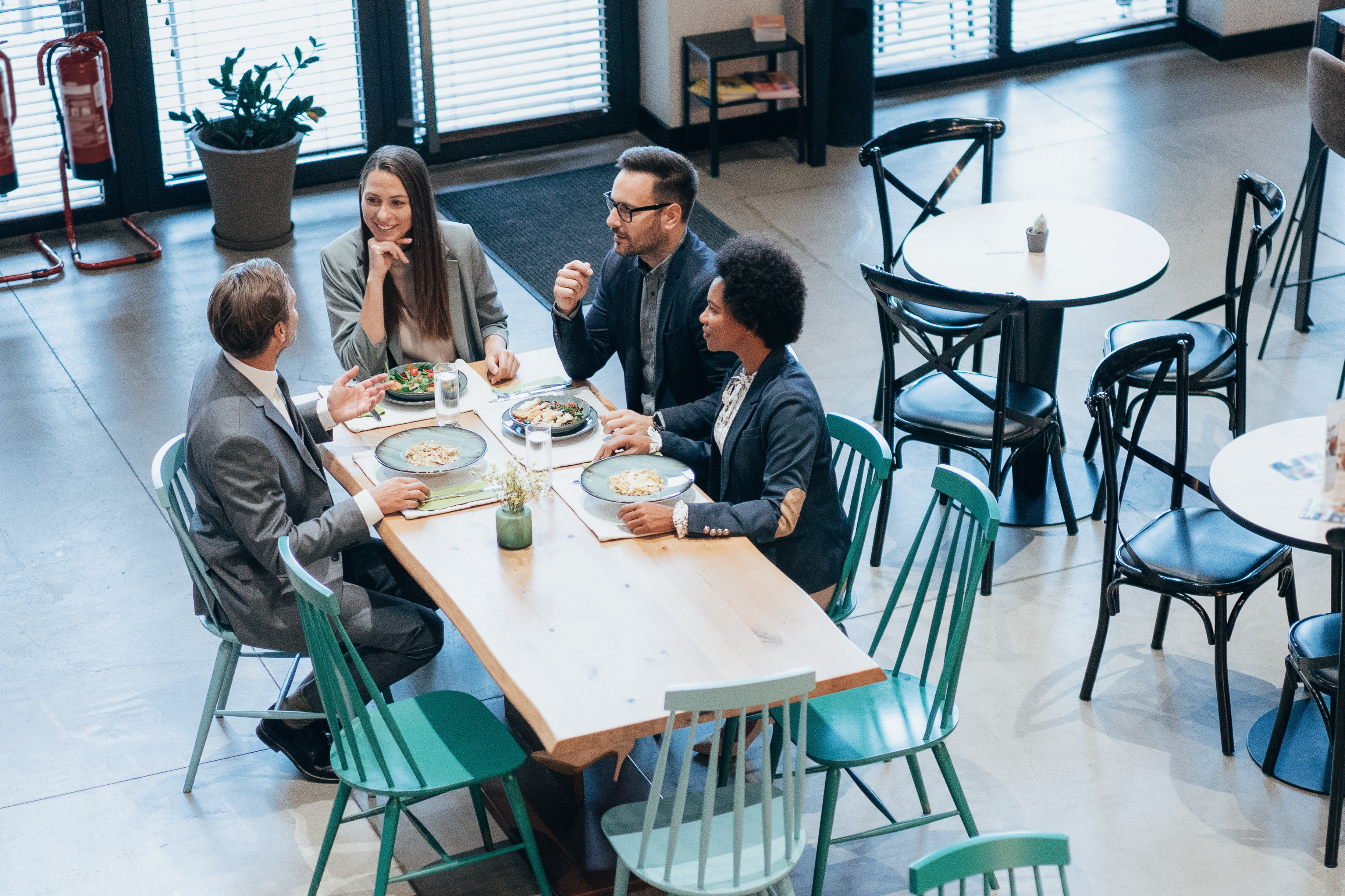 A group of four colleagues, two men and two women, sit eating and talking at a table in a casual restaurant with natural light from large windows