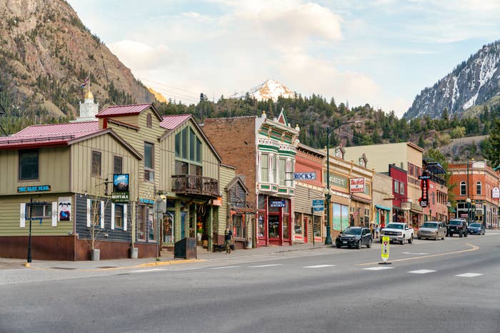 A quaint mountain town features a row of colorful storefronts and shops along a street with parked cars, with a snow-capped mountain in the background