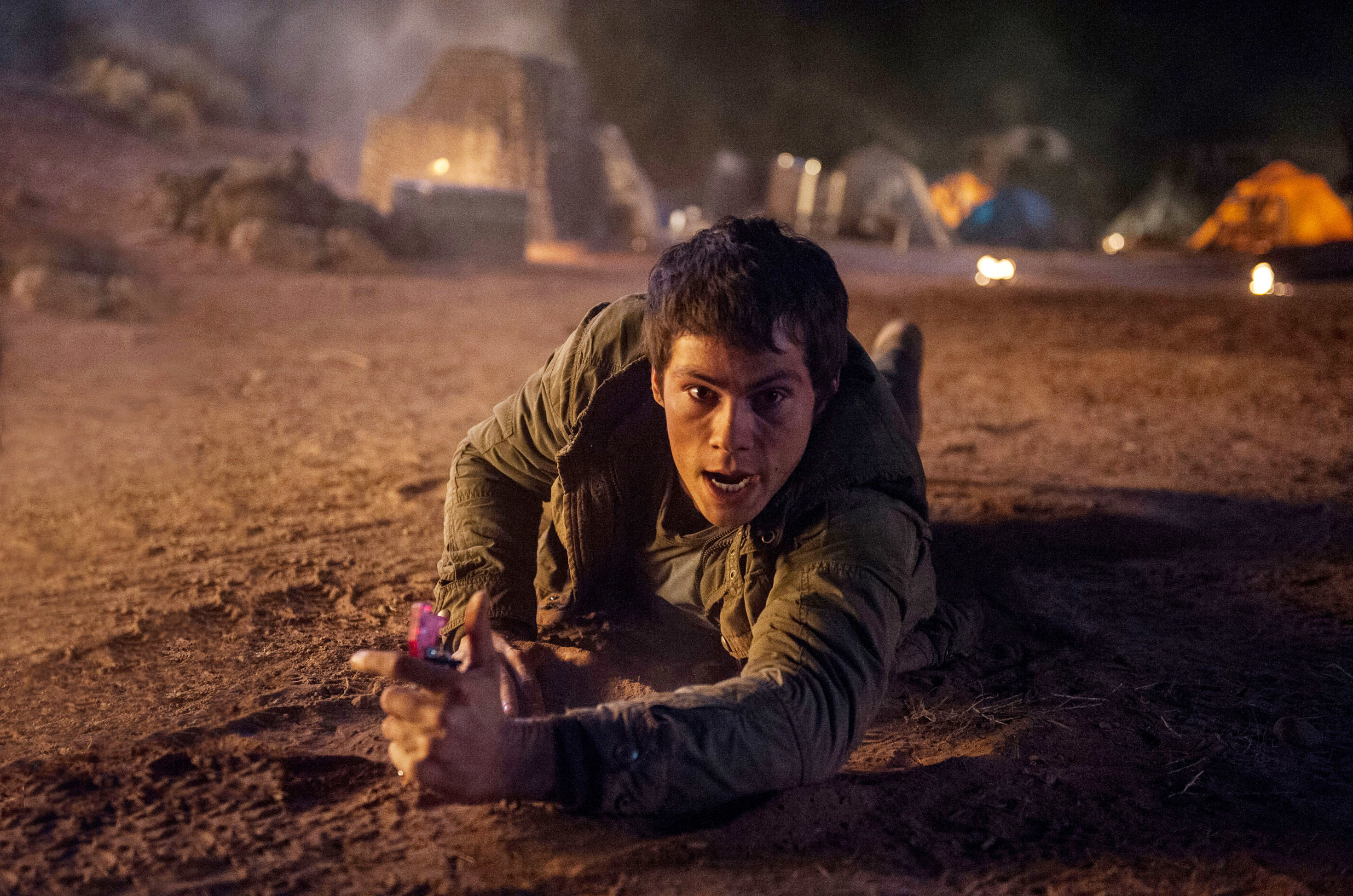 Dylan O'Brien, in rugged clothing, crawls urgently on the ground with a determined expression, holding a small device, in an intense scene
