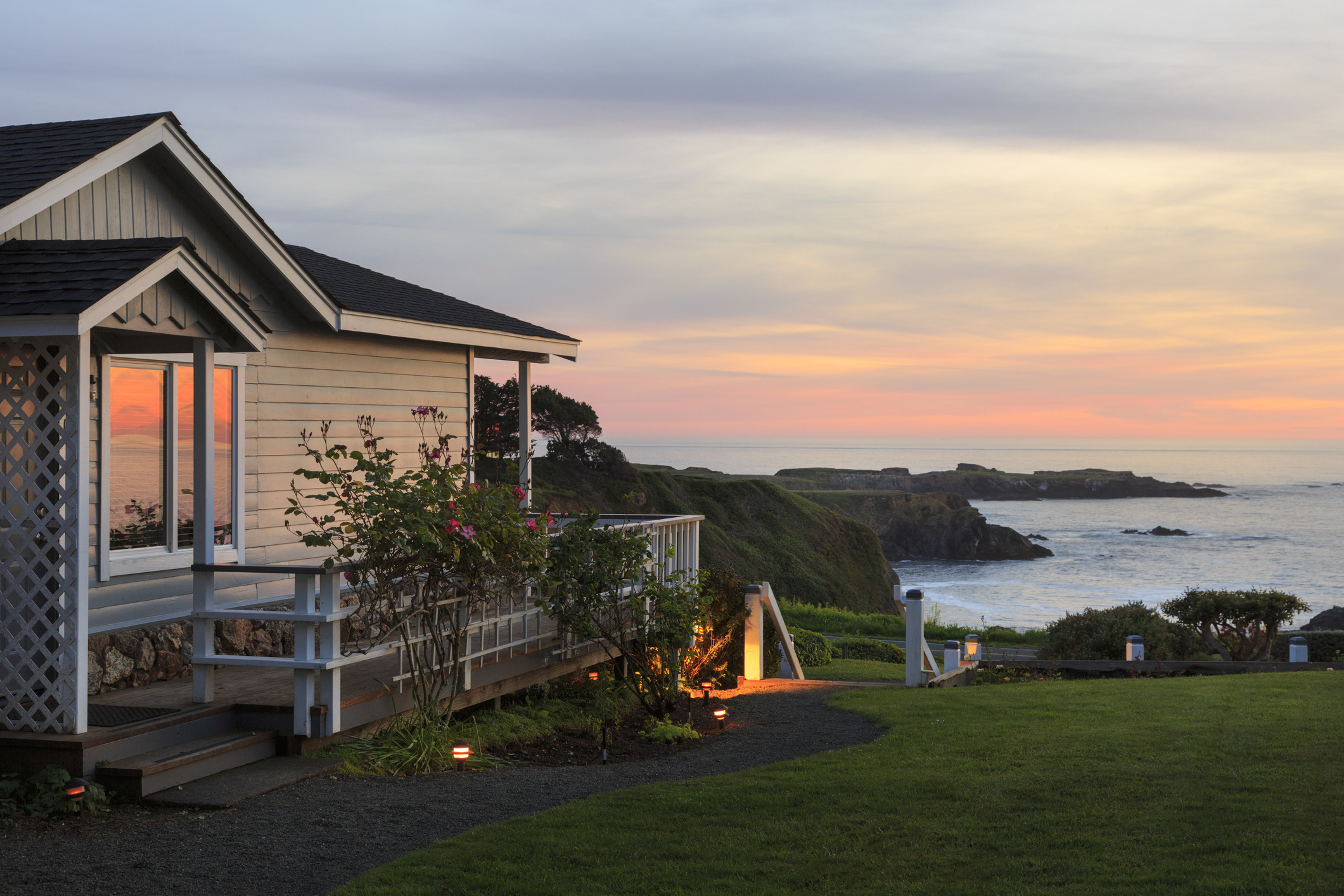A cozy house with a garden overlooks the ocean at sunset, with cliffs and the sea visible in the distance