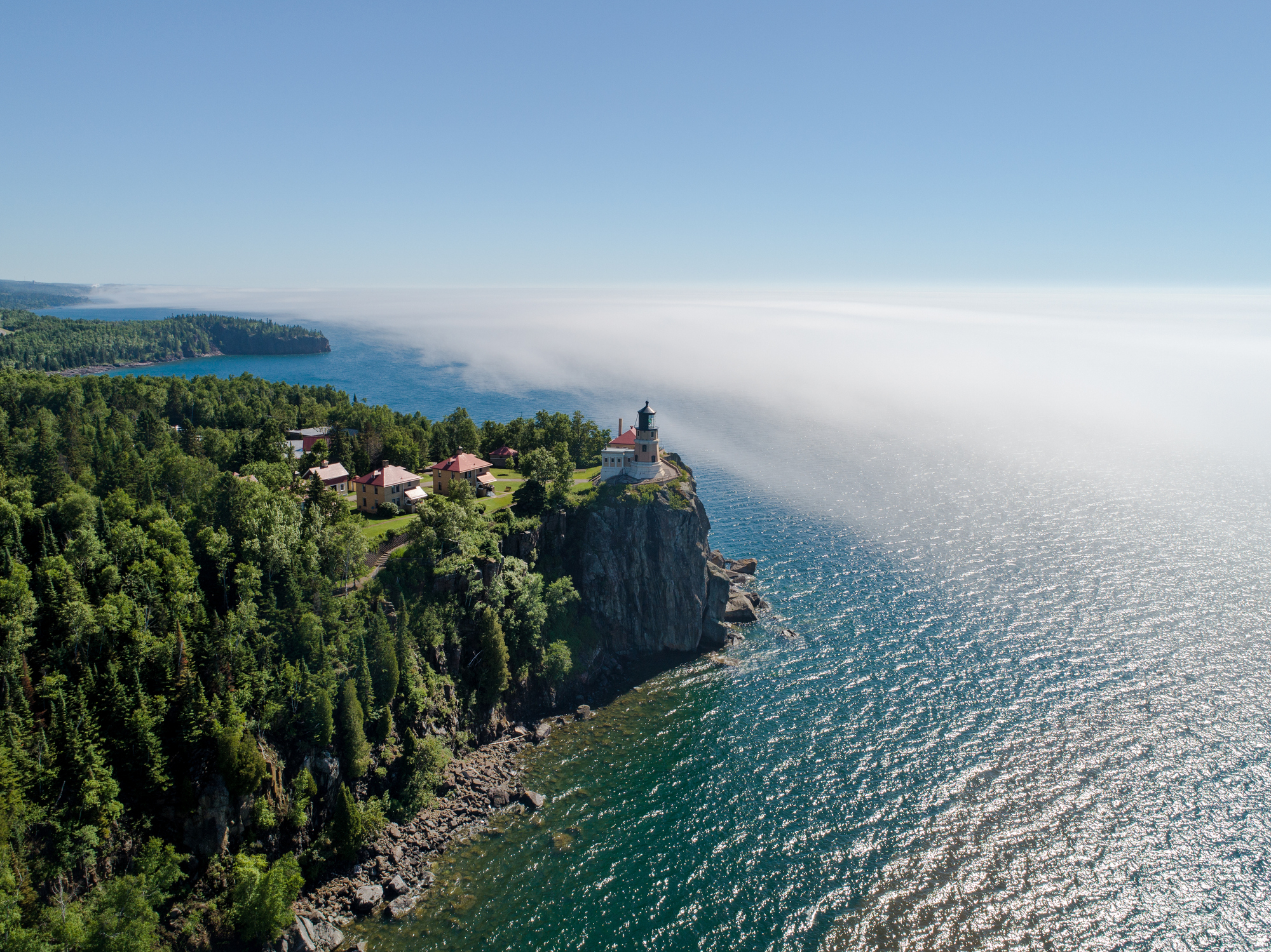 Aerial view of Split Rock Lighthouse perched on a cliff, overlooking a vast body of water and surrounded by lush greenery and forested landscape