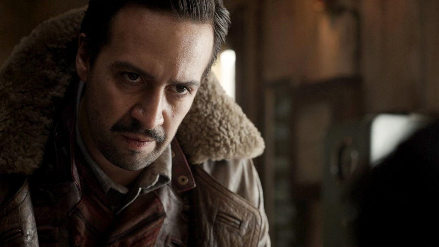 Lin-Manuel Miranda in a serious expression, wearing a jacket with a shearling collar, in a scene from a TV show or movie