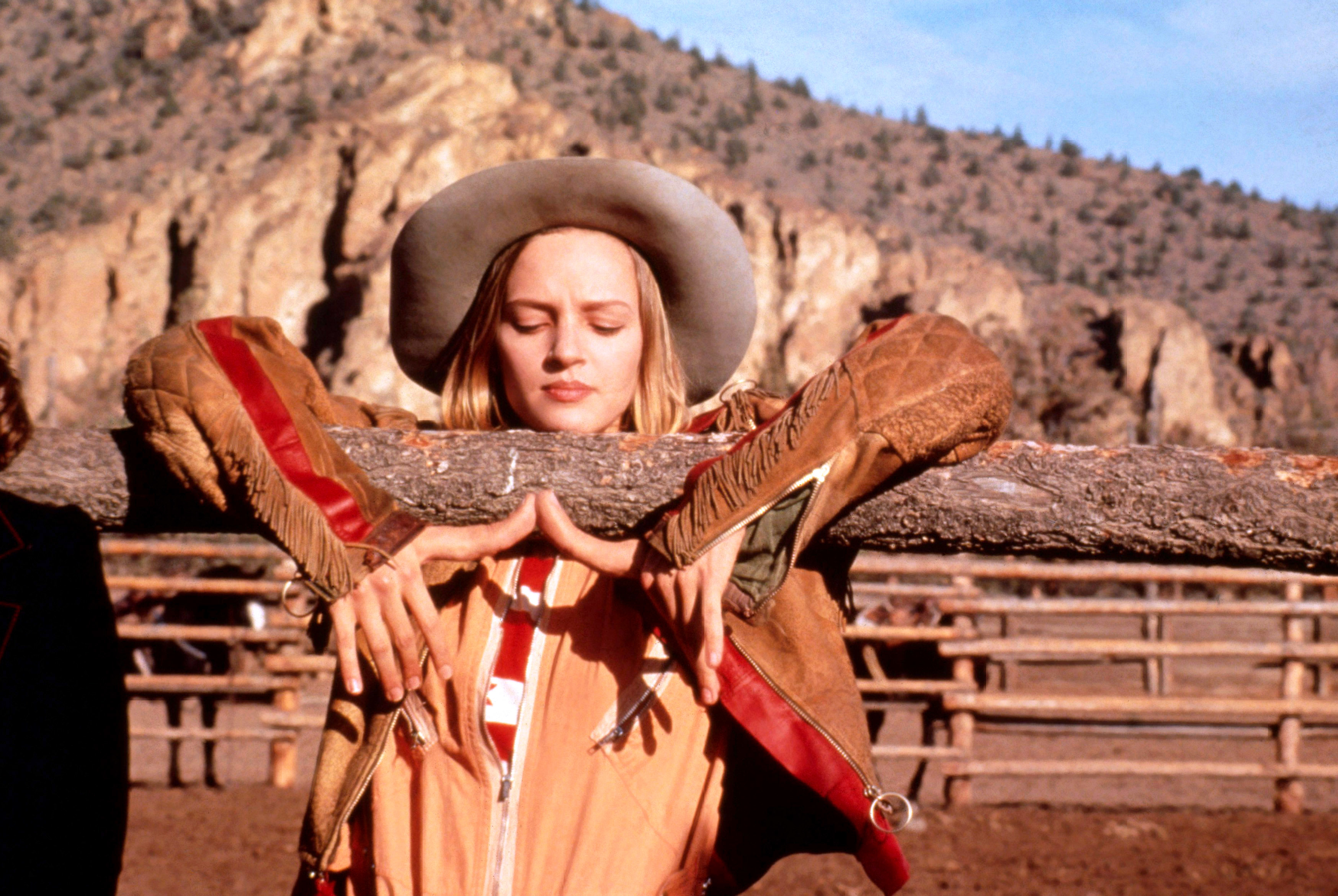 Uma Thurman leans on a wooden fence in a western-themed outfit with a cowboy hat, in a ranch setting with mountains in the background