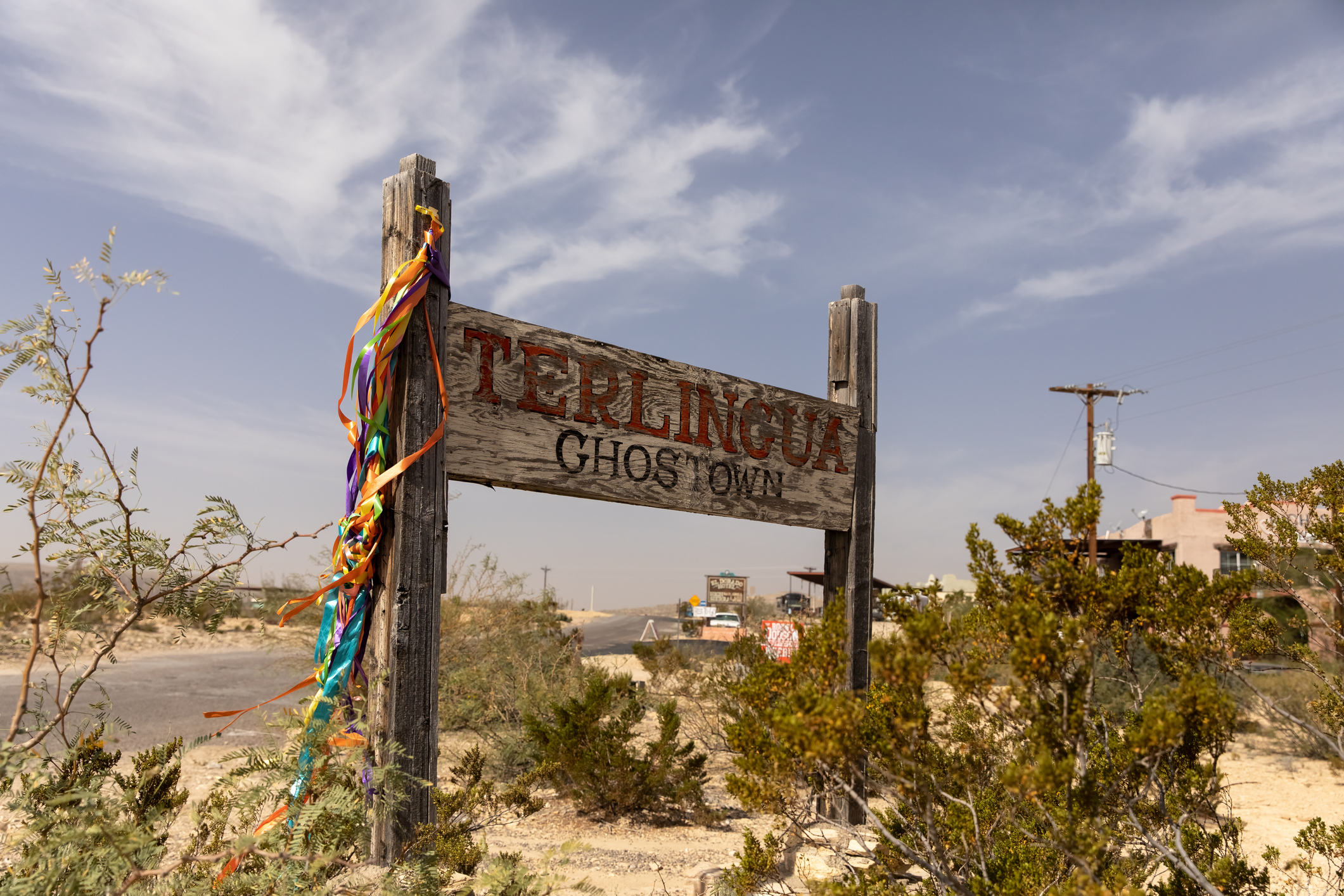 Sign reads "Terlingua Ghost Town" adorned with colorful ribbons, set in a dry, desert landscape with sparse vegetation