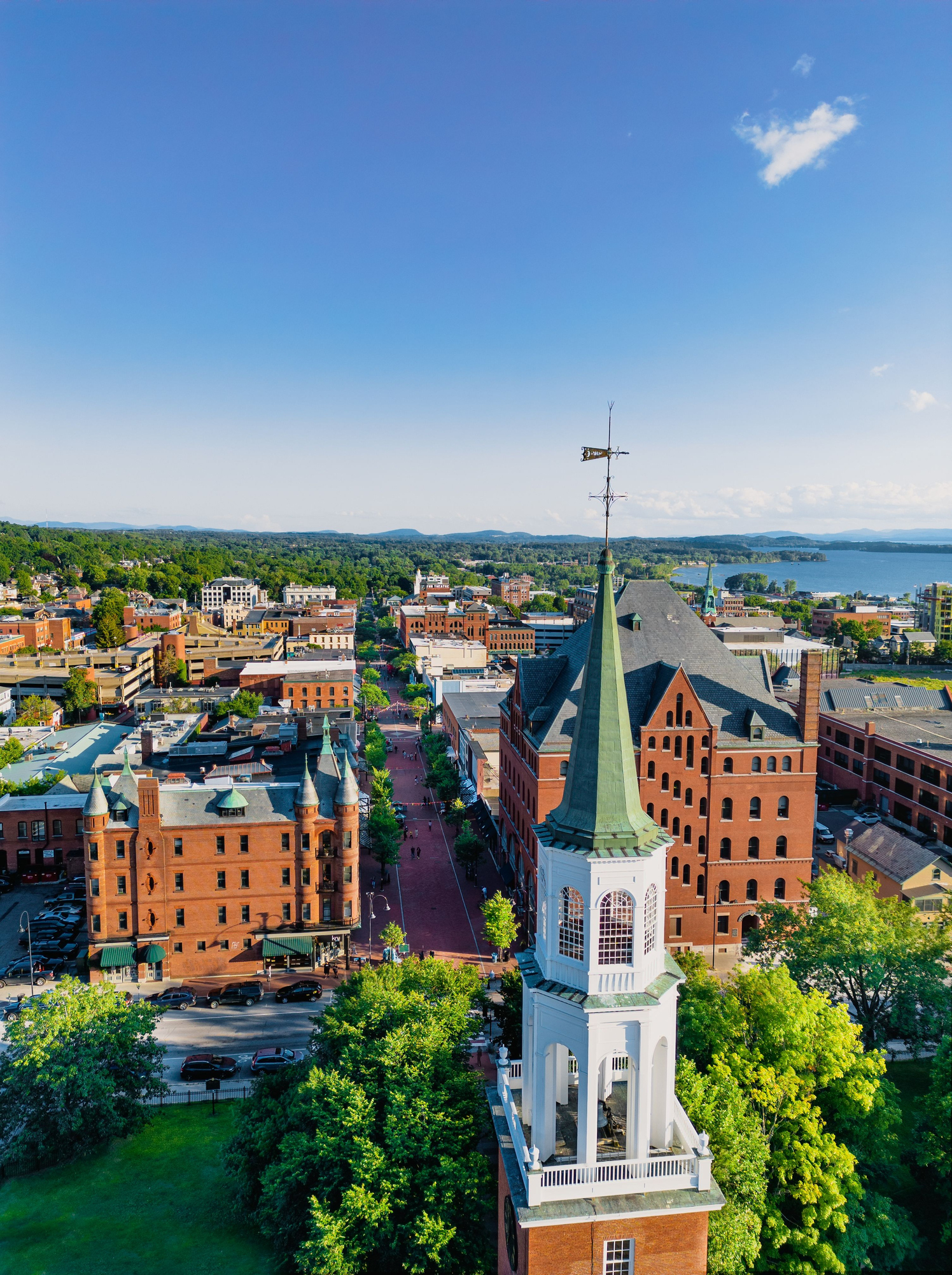 Aerial view of a coastal town with a prominent white clock tower in the foreground and buildings, trees, and distant water visible in the background