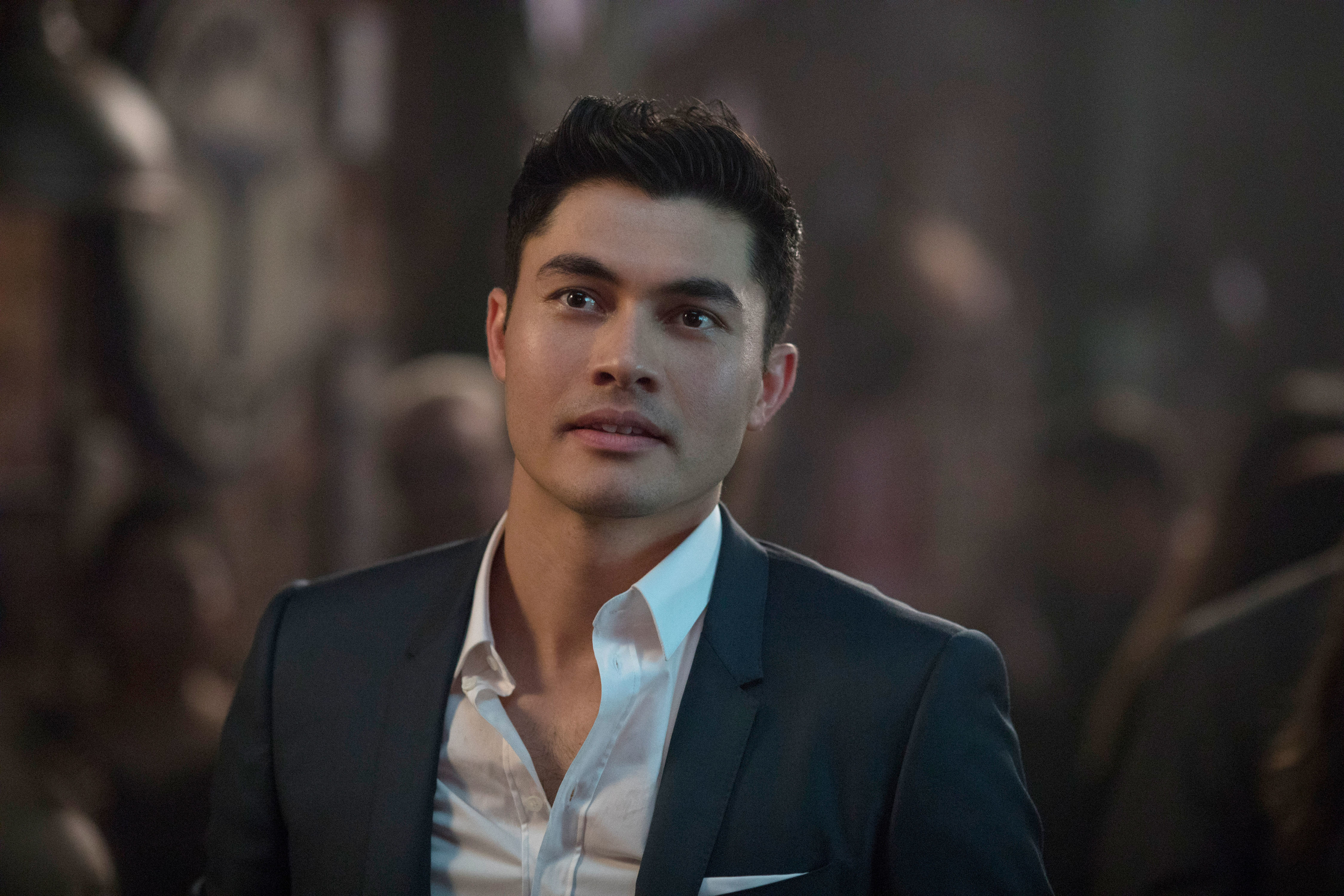 Henry Golding dressed in a dark suit with a white shirt, standing in a dimly lit environment