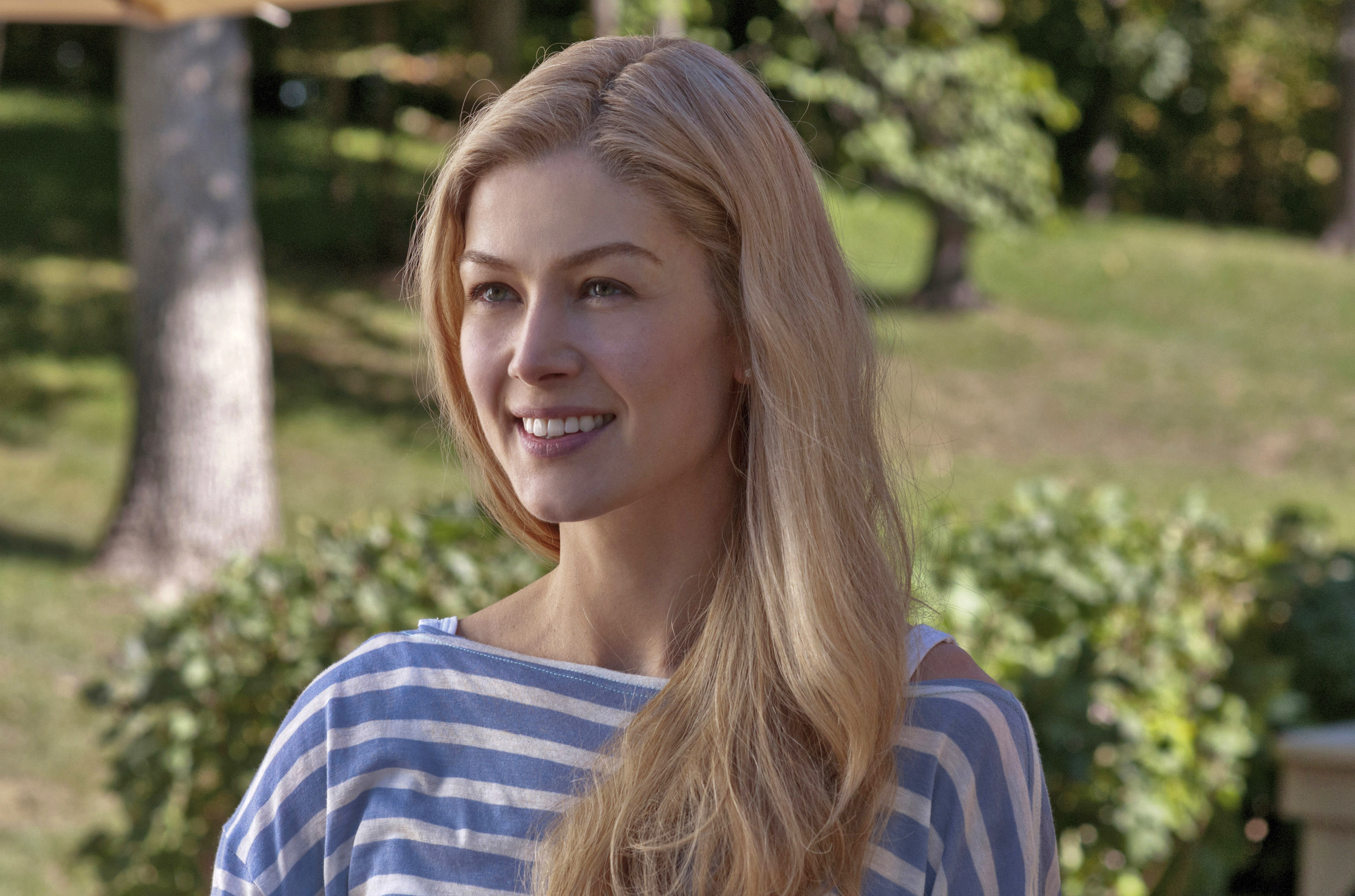 Rosamund Pike, in a casual striped top, stands outside with a smile. Trees and greenery are visible in the background