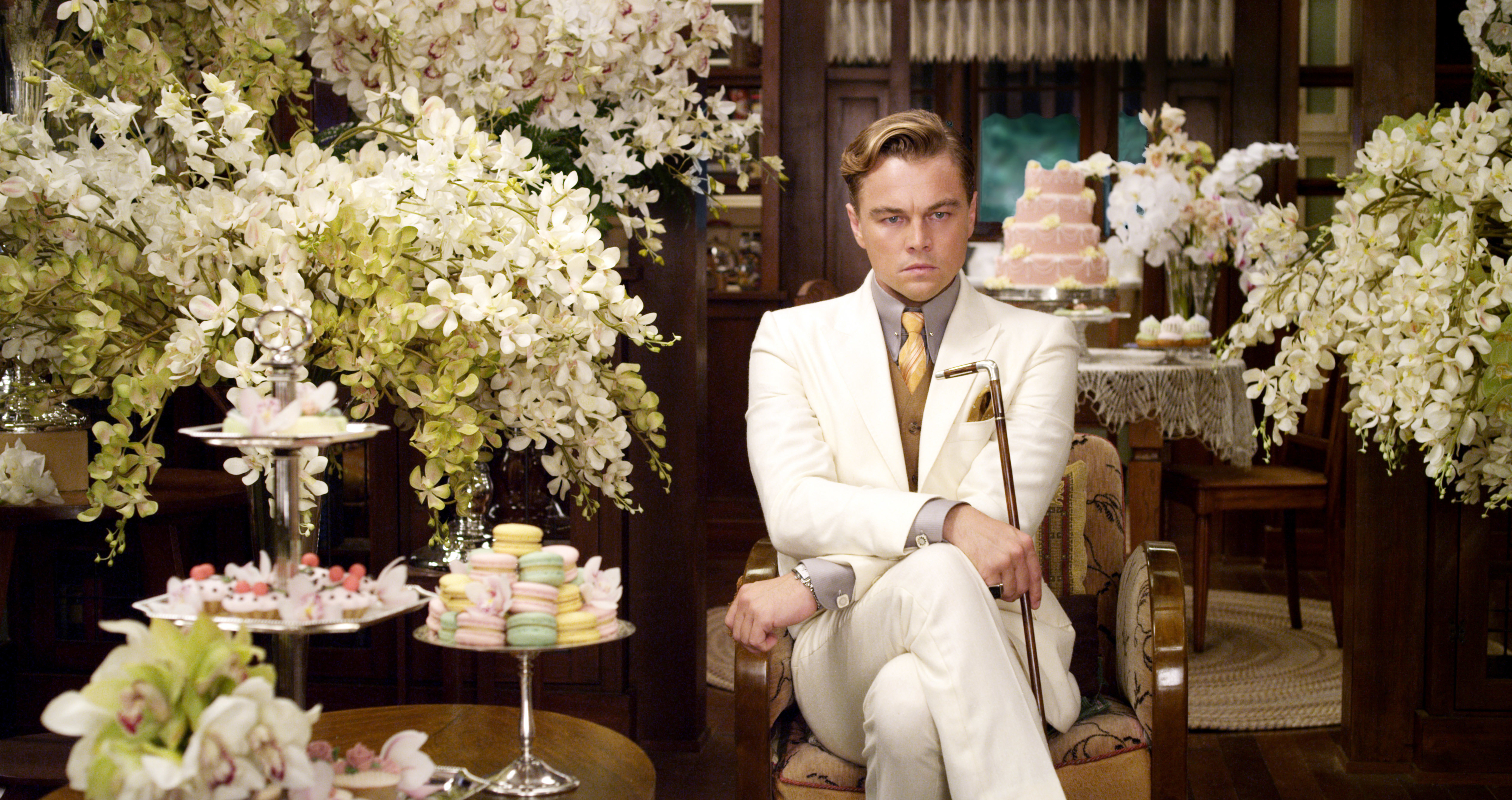 Leonardo DiCaprio as Jay Gatsby sits in a luxurious setting with floral decorations, desserts, and a lavish cake in a scene from &quot;The Great Gatsby.&quot;