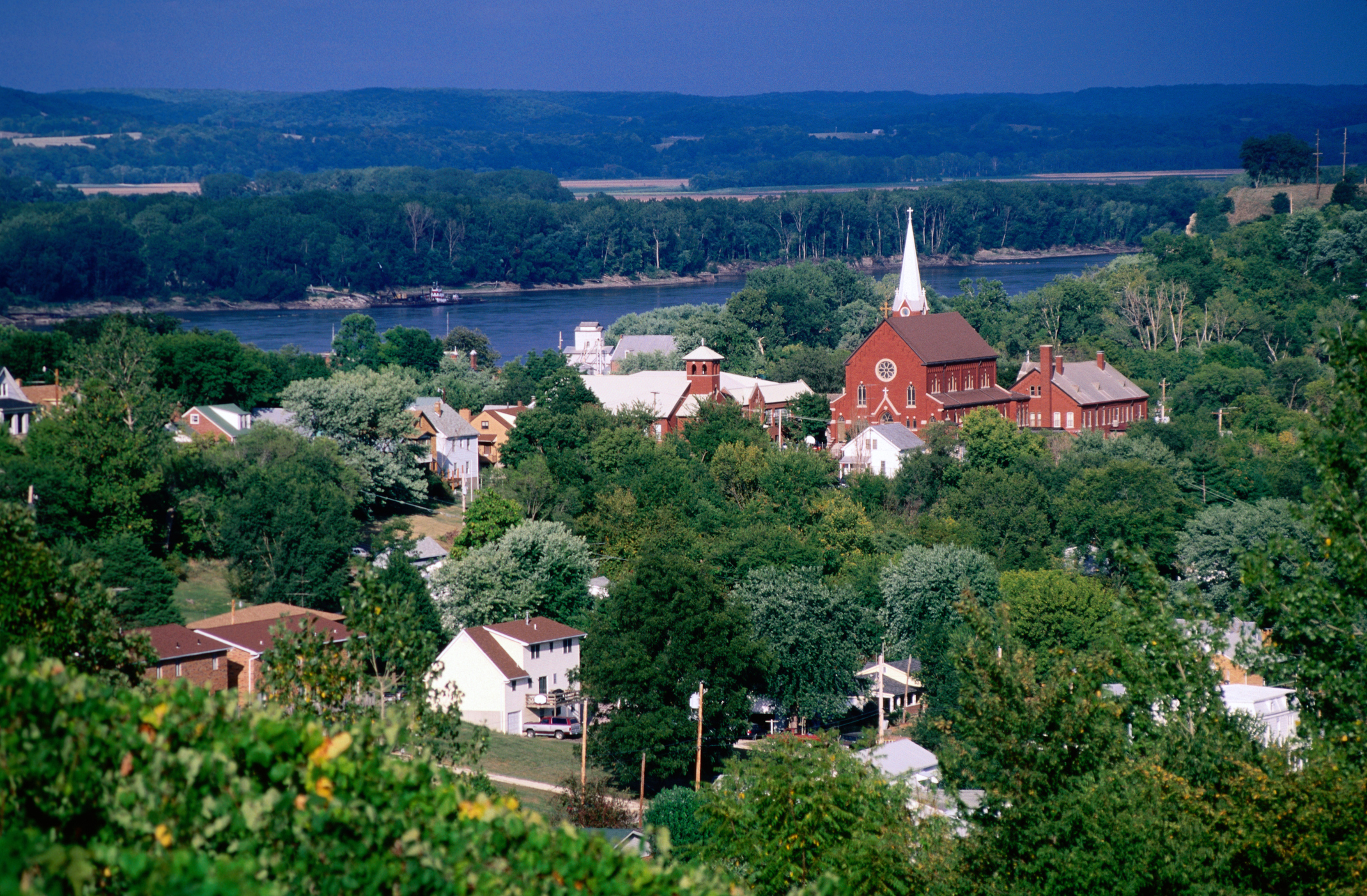 A picturesque landscape showing a small town with a river in the background, surrounded by lush green trees and a prominent red church with a white steeple