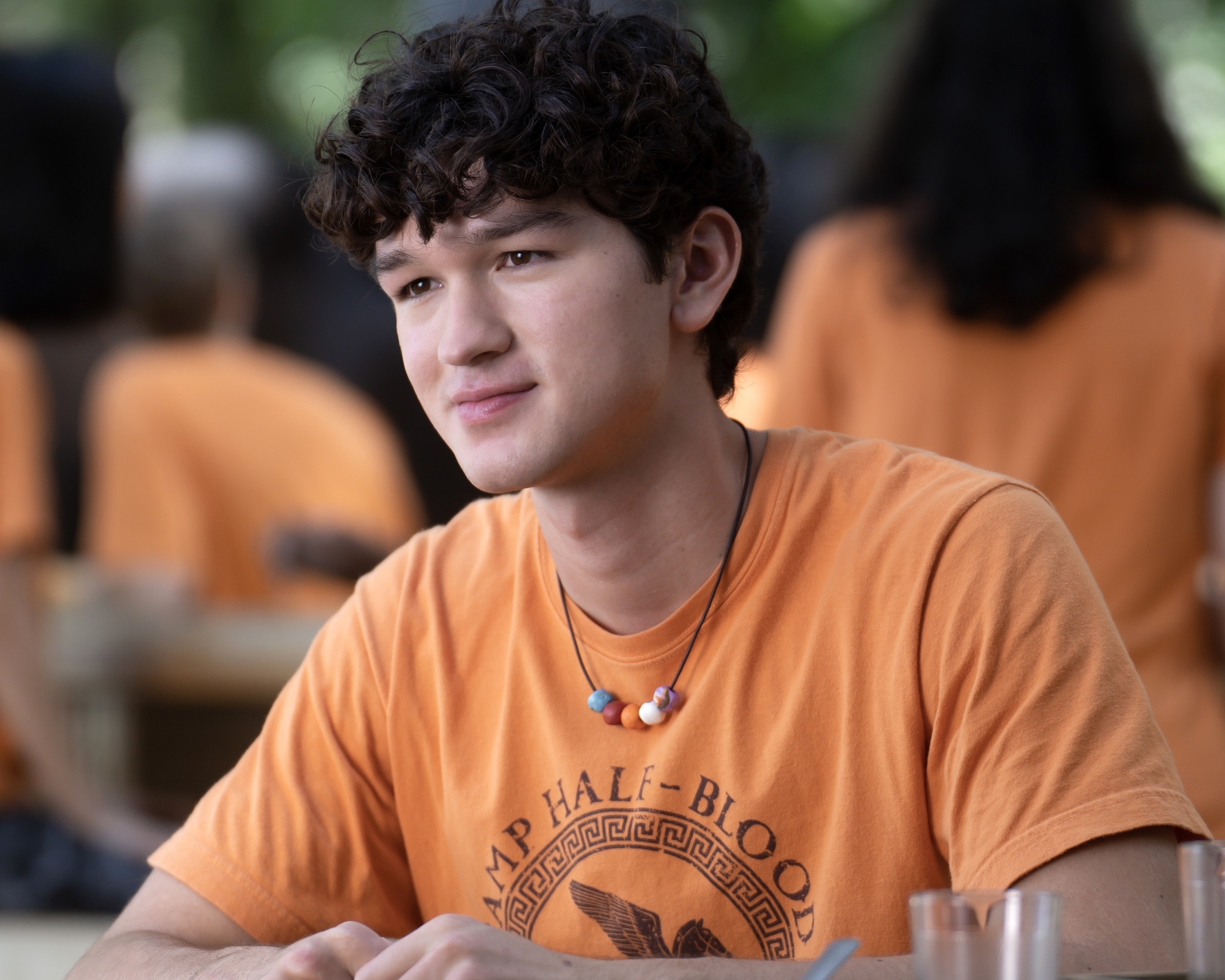Charlie Bushnell sits at a table, wearing an orange &quot;Camp Half-Blood&quot; t-shirt and a beaded necklace