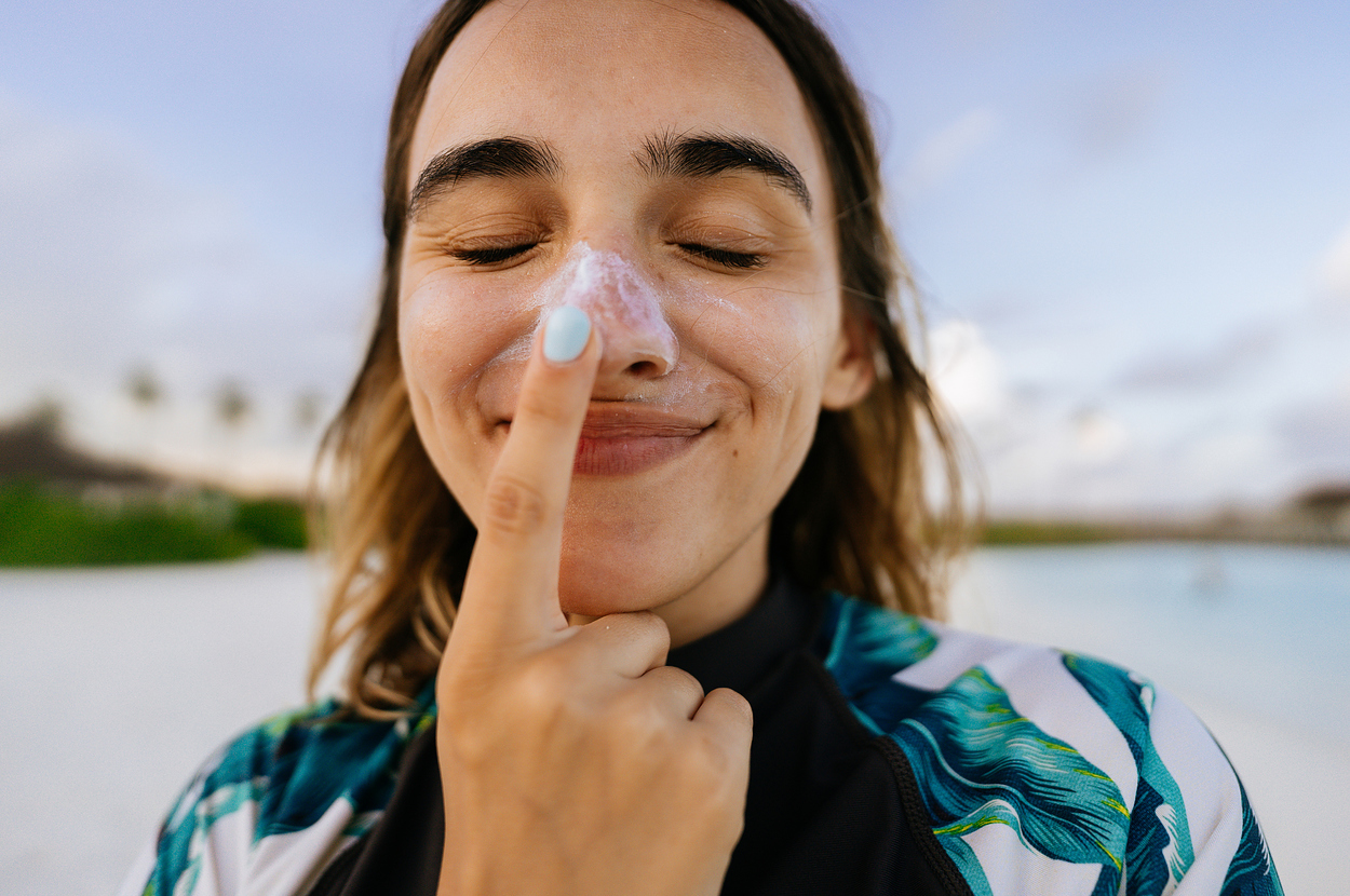 Person smiling, applying sunscreen on their nose with a finger, standing outside with a beach background