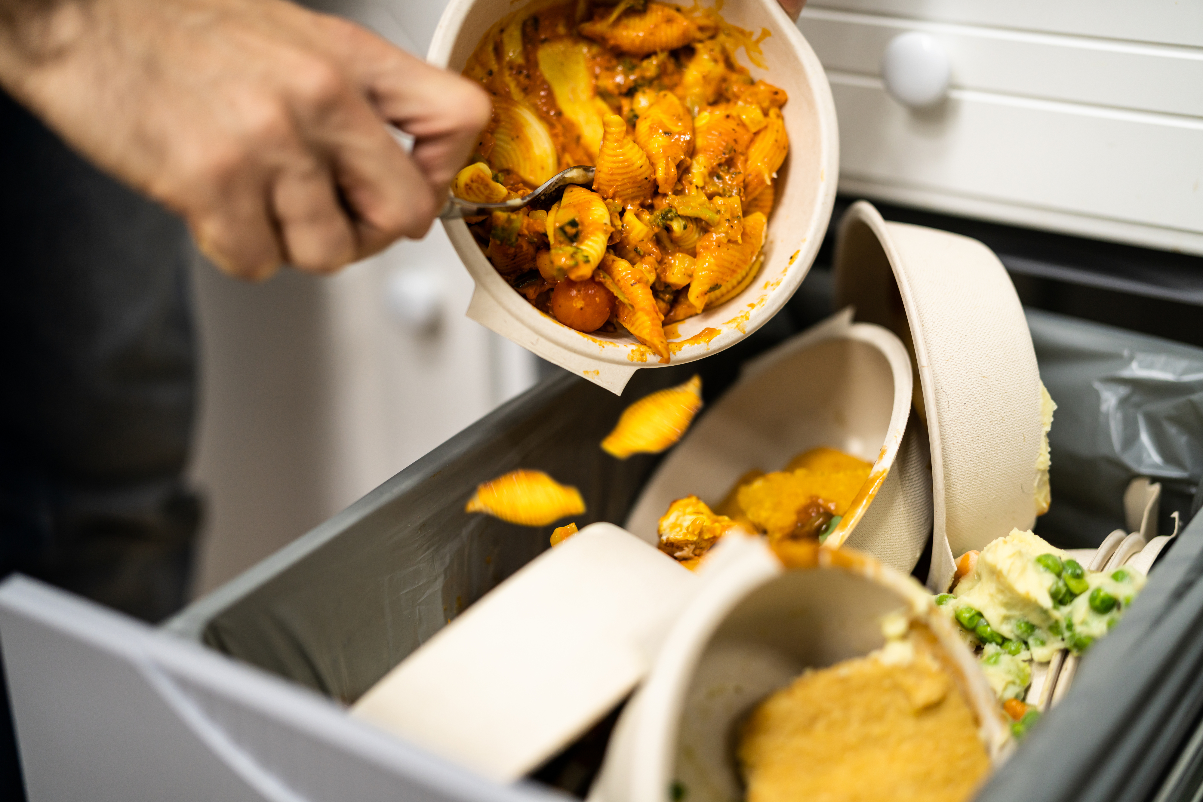A person's hand is scraping leftovers from a bowl into a trash bin, visibly illustrating food waste, which is a concern in many workplaces