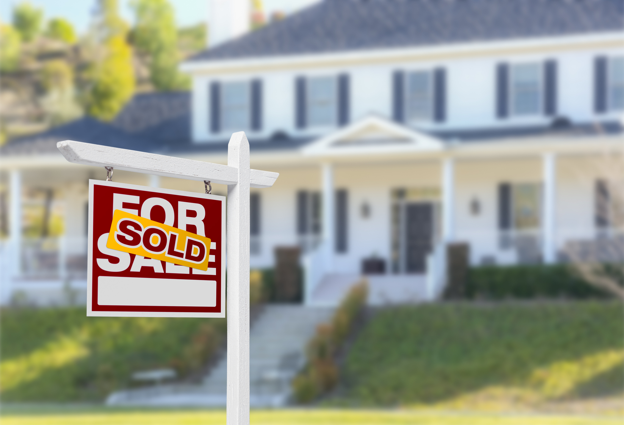 A &quot;For Sale&quot; sign with a &quot;Sold&quot; sticker in front of a large house, indicating the house has been sold