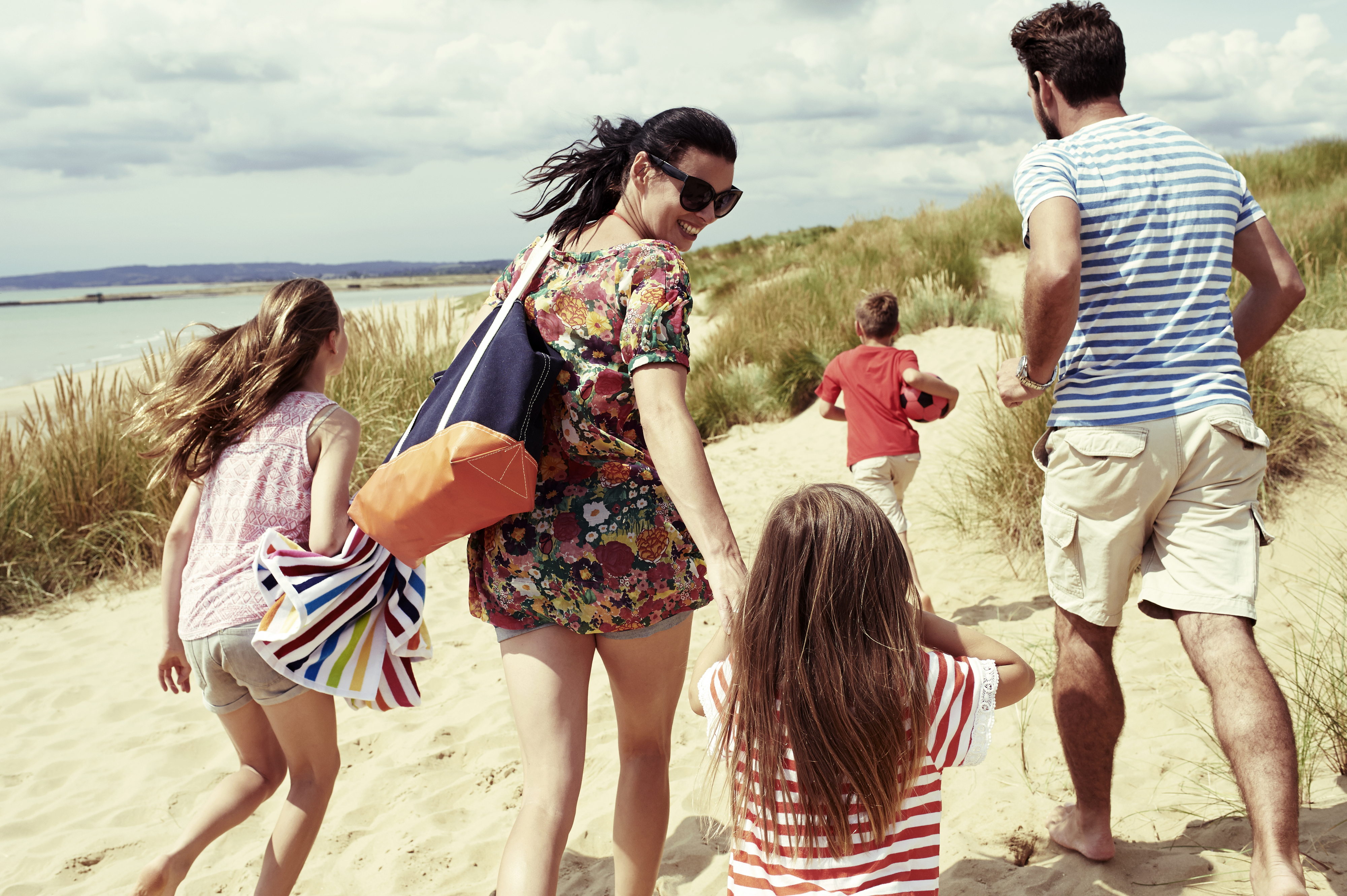 A family of five walks on a sandy beach. Adults and children carry bags and wear casual, summery clothes. The adults are holding the hands of the younger children