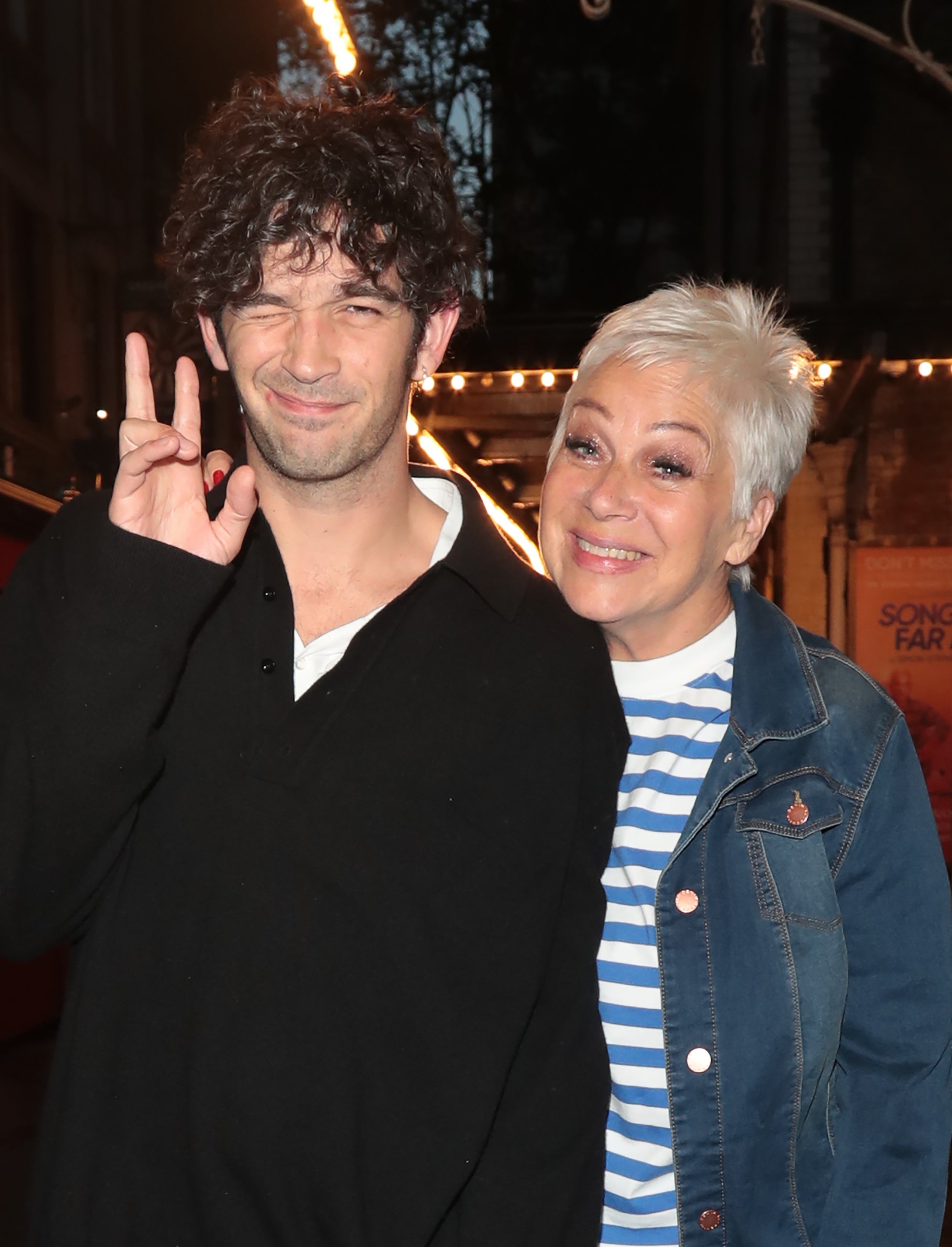 Matty Healy and Denise Welch stand together outdoors at night, posing for a photo. Matty Healy wears a black outfit, while Denise Welch is in a denim jacket and striped shirt