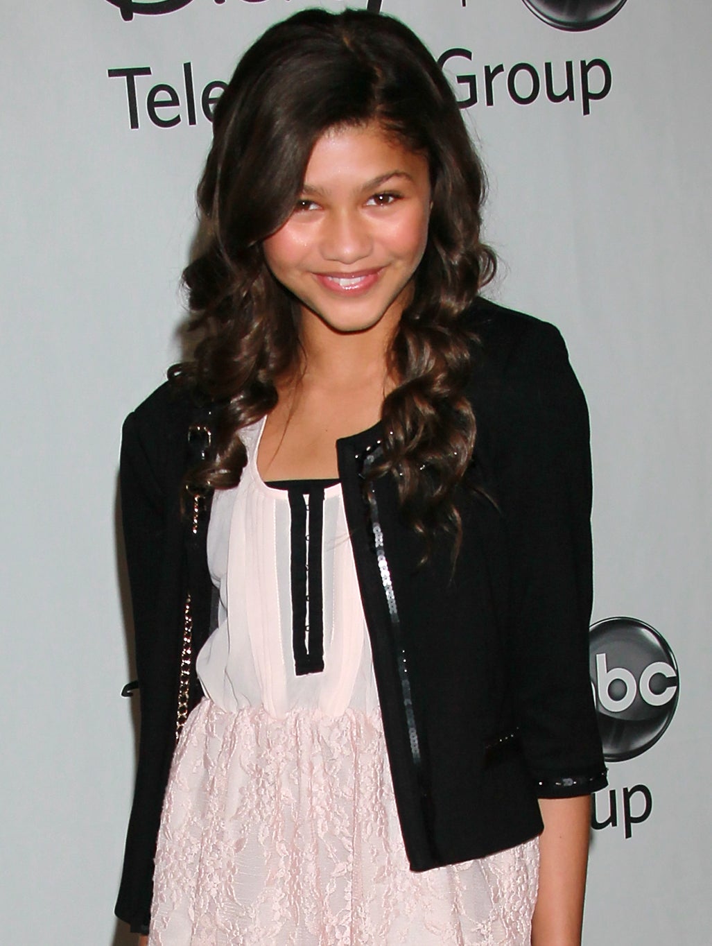 Zendaya poses on the red carpet wearing a black jacket, a pale pink dress, patterned tights, and black boots at a Disney/ABC Television Group event