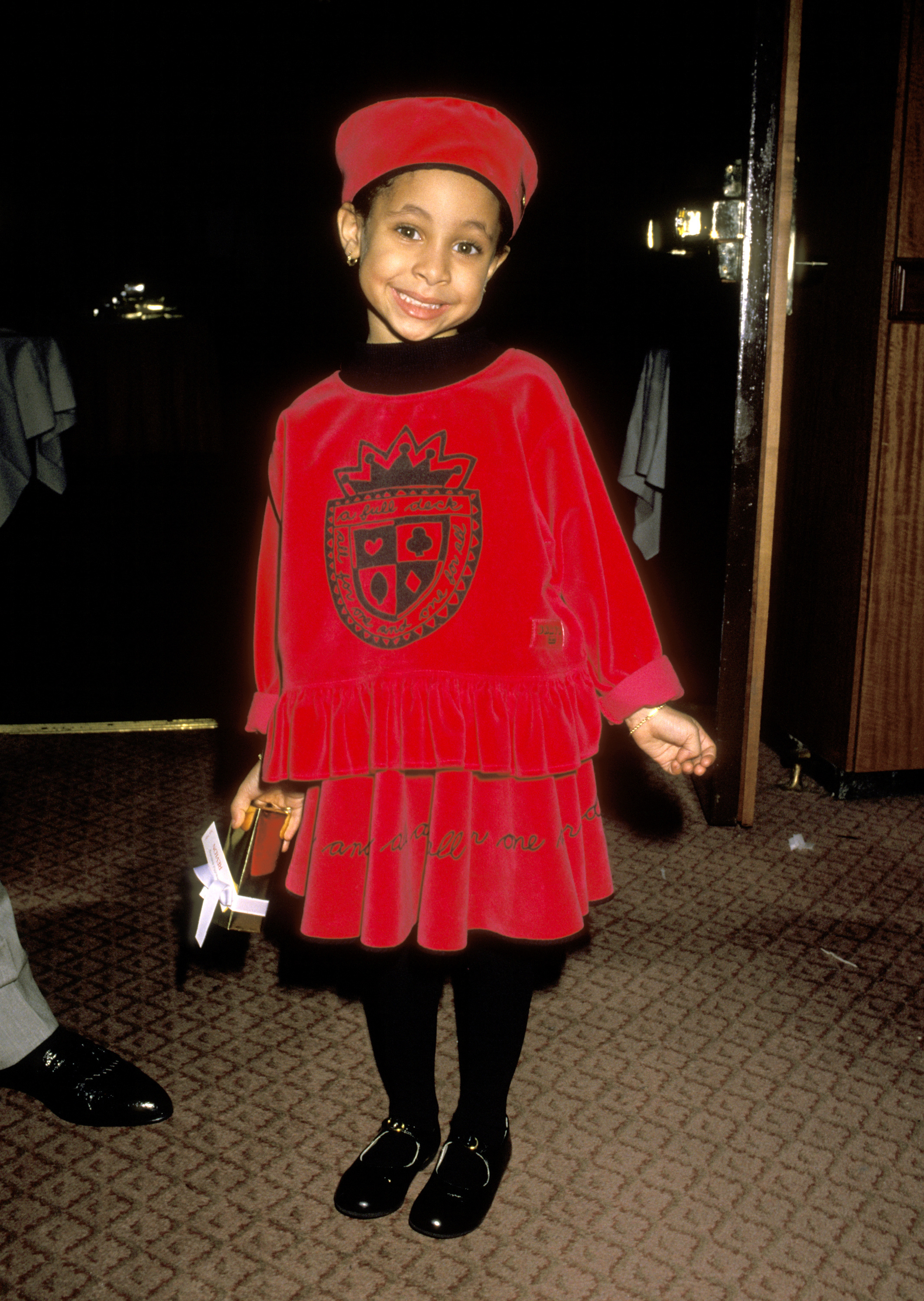 A young Raven-Symoné is smiling, wearing a red beret and a matching velvet dress with a crest emblem, holding a toy, standing indoors on a patterned carpet