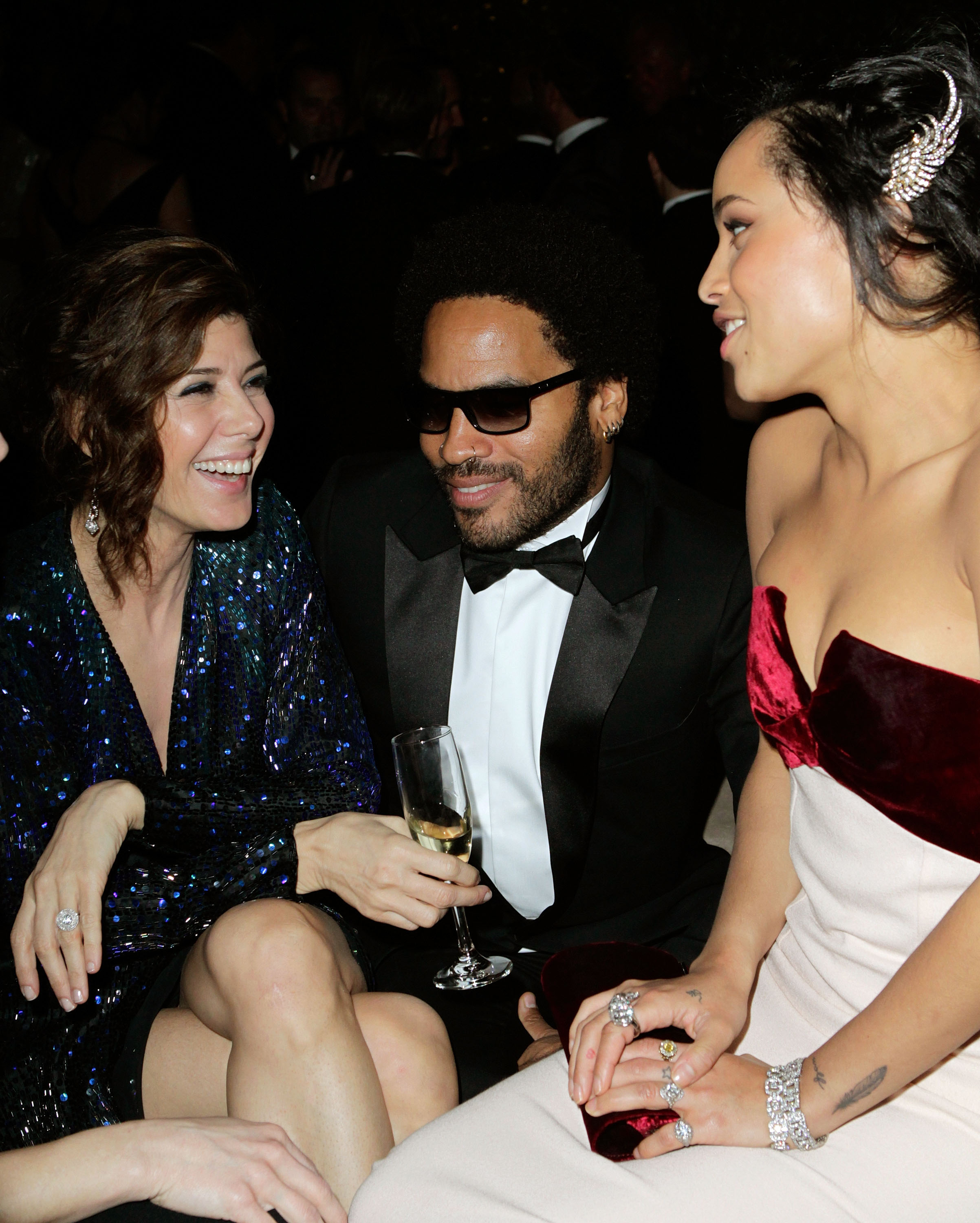 Marisa Tomei in a sequined dress, Lenny Kravitz in a tuxedo with sunglasses, and Zoë Kravitz in a strapless dress with red accents, seated and smiling at an event