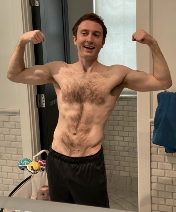 Daryl Sabara flexing muscles in front of a mirror, shirtless, wearing black shorts in a bathroom. Baby's head visible at the bottom of the image. Names not provided