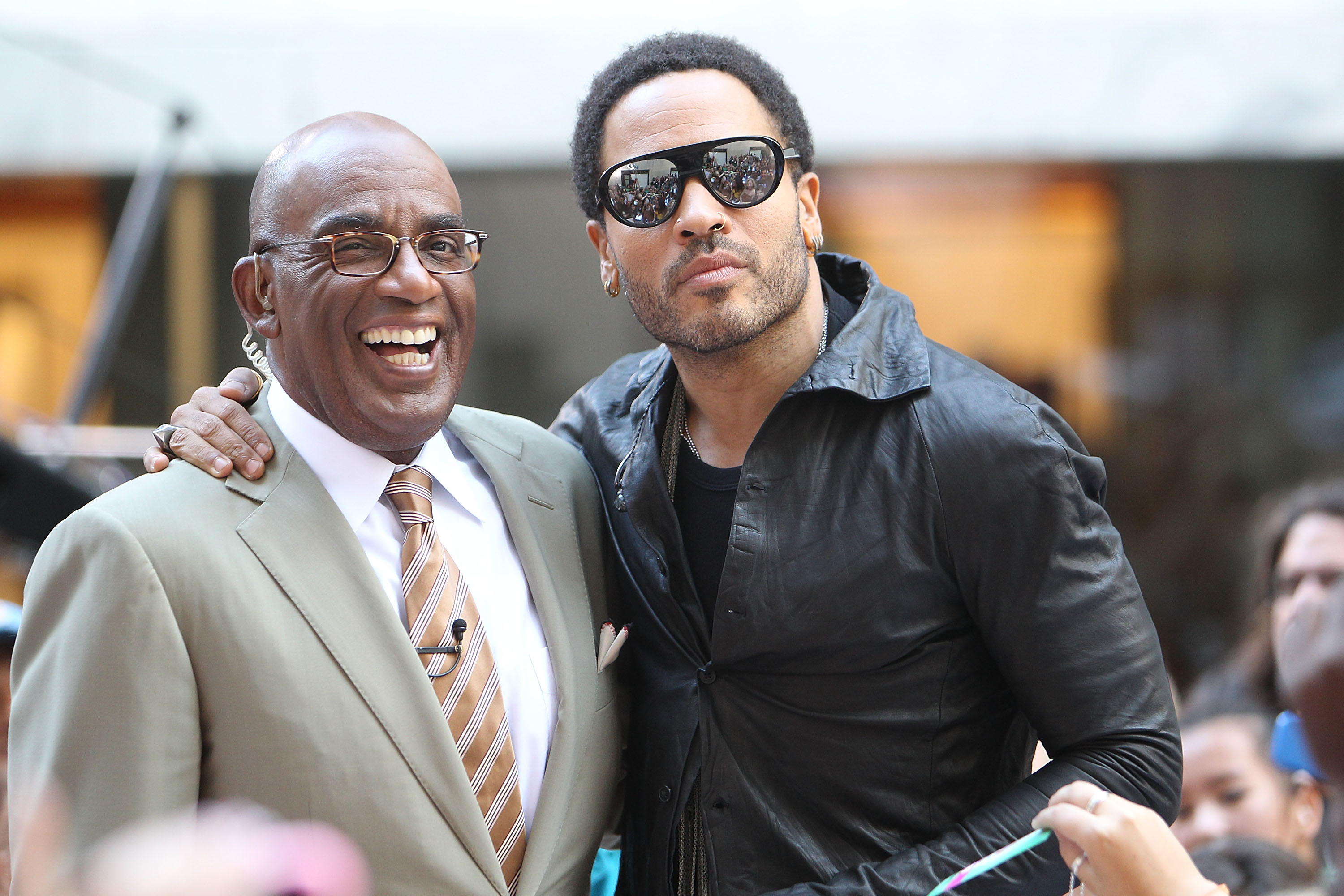 Al Roker in a tan suit and tie smiles while standing next to Lenny Kravitz, who is wearing a black leather jacket and sunglasses