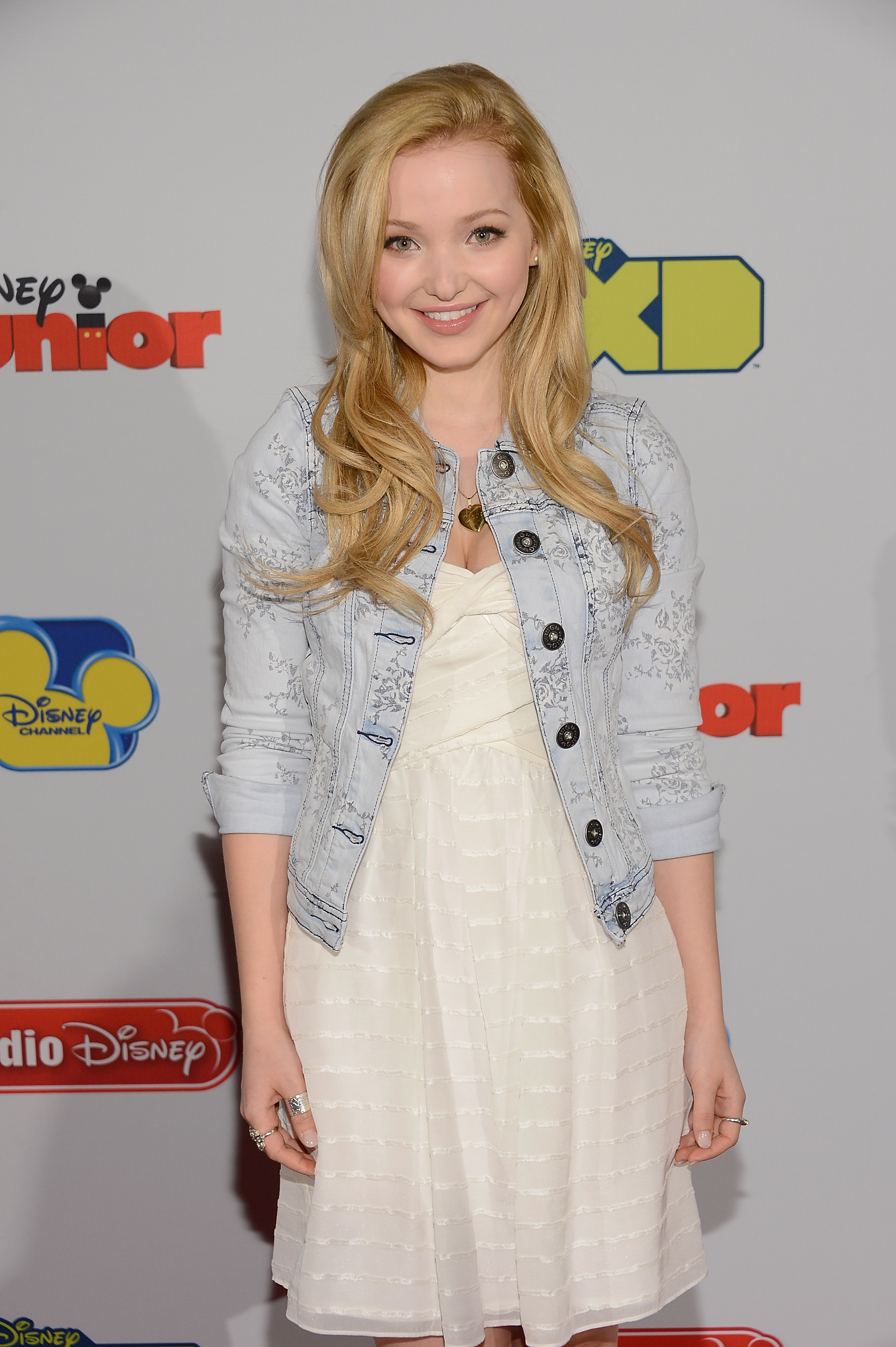 Dove Cameron at a Disney event, wearing a white dress and light denim jacket, smiling at the camera