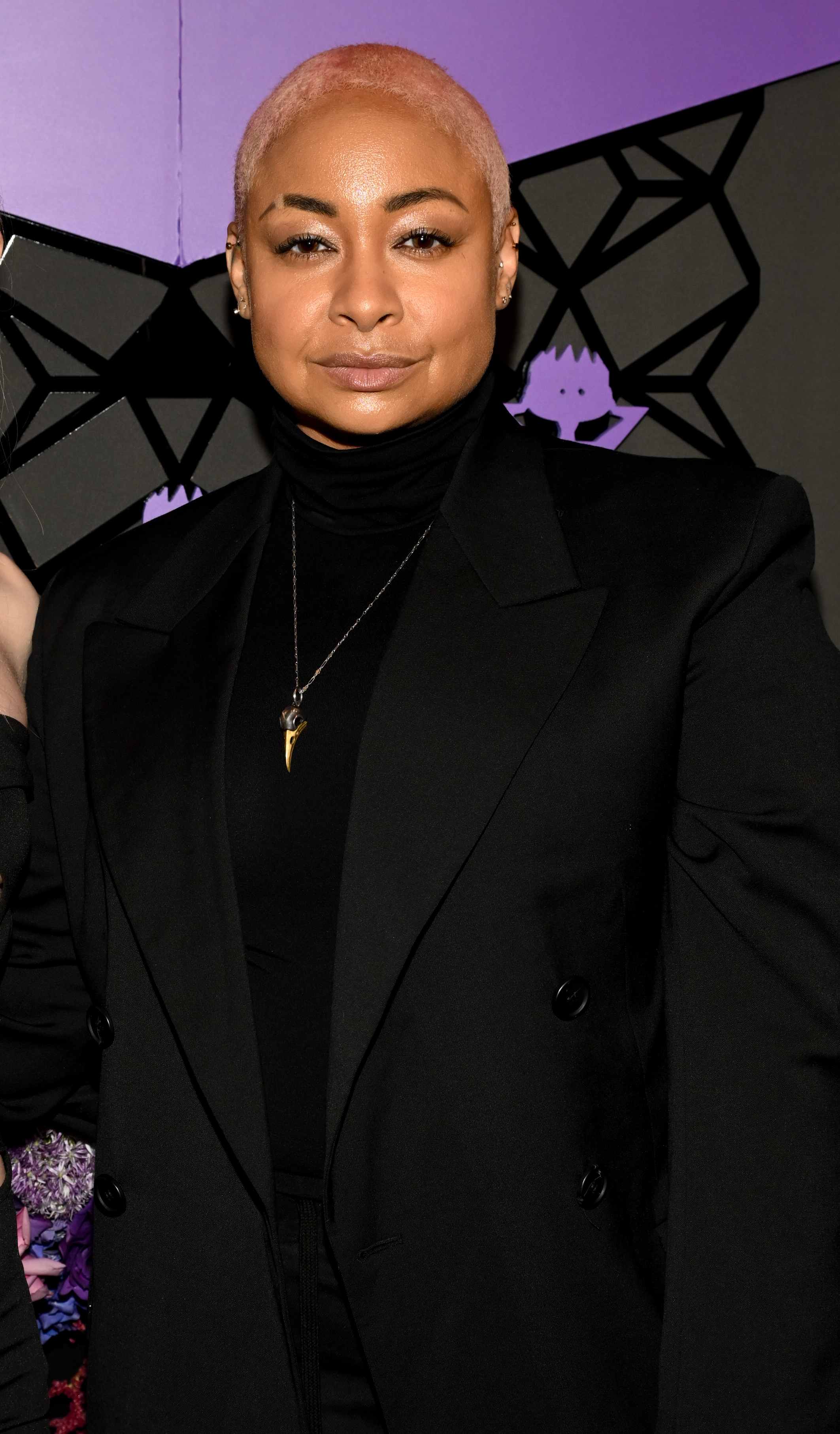 Raven-Symoné on a purple and black background, wearing a black double-breasted blazer, black turtleneck, and a necklace with a gold pendant