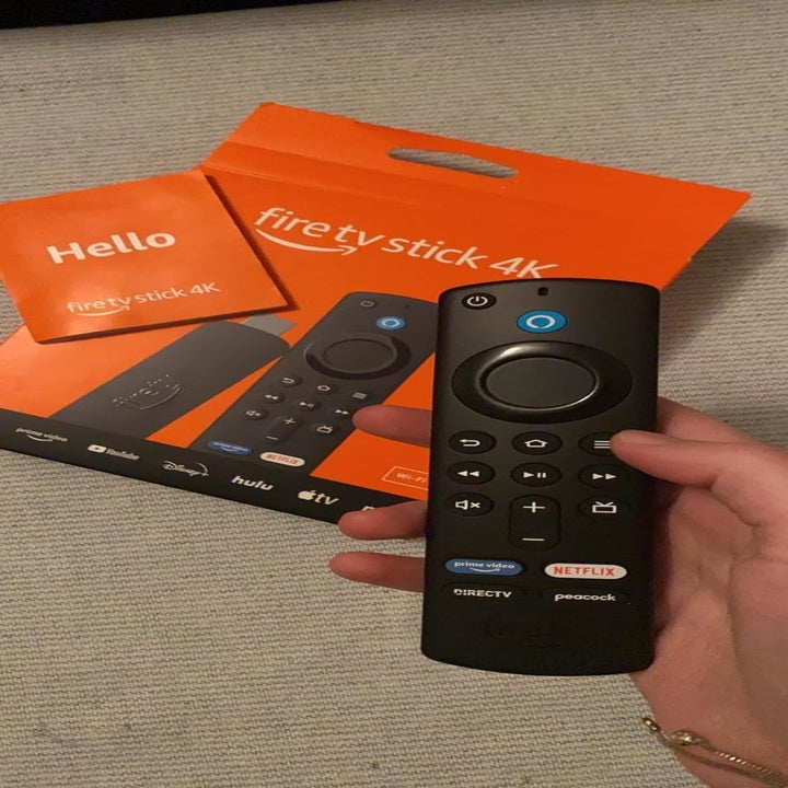 A hand holding an Amazon Fire TV Stick 4K remote, with the orange Fire TV Stick 4K box and setup guide visible in the background
