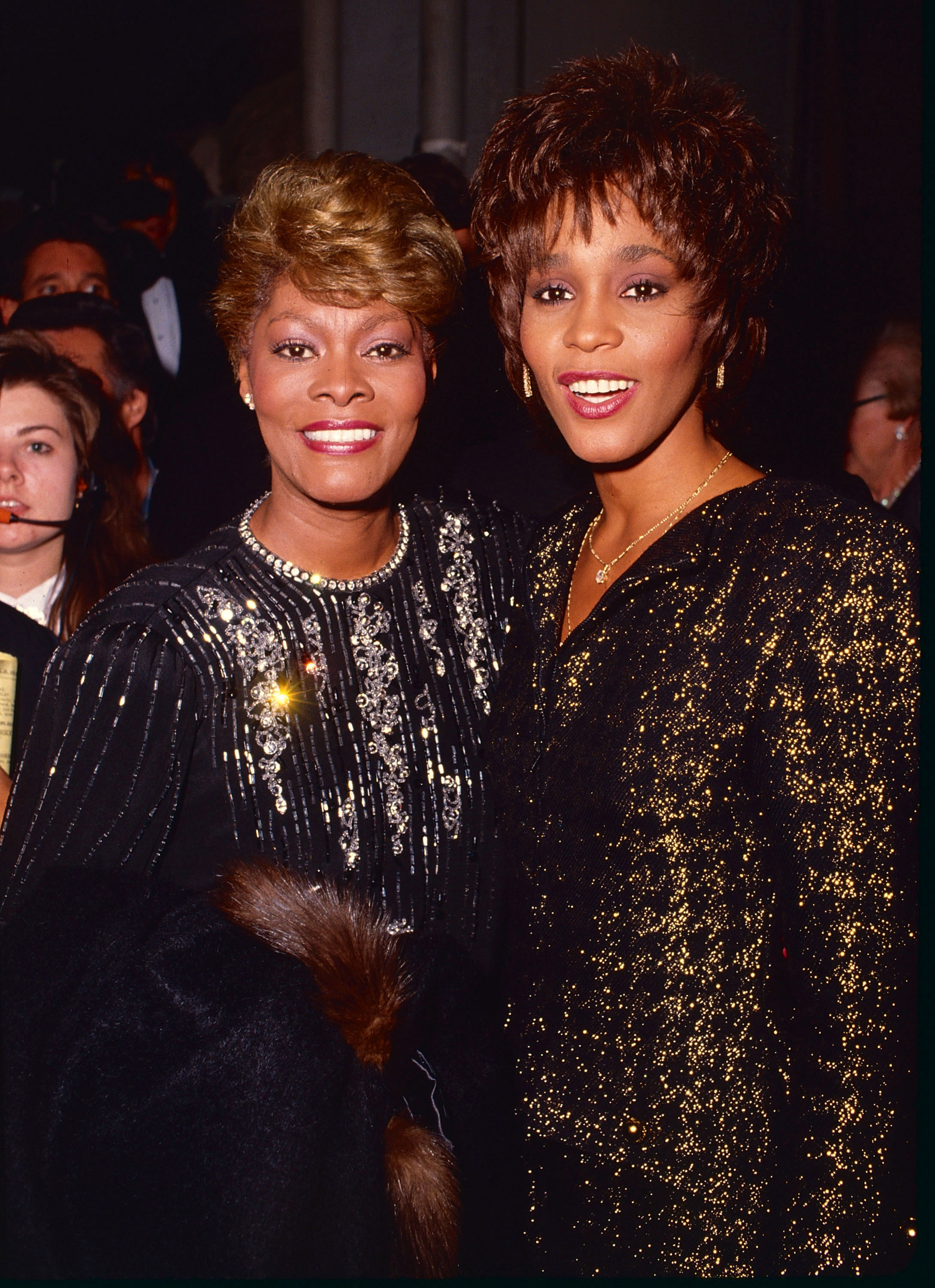 Dionne Warwick and Whitney Houston pose together at a formal event, both in elegant black gowns with sparkling embellishments
