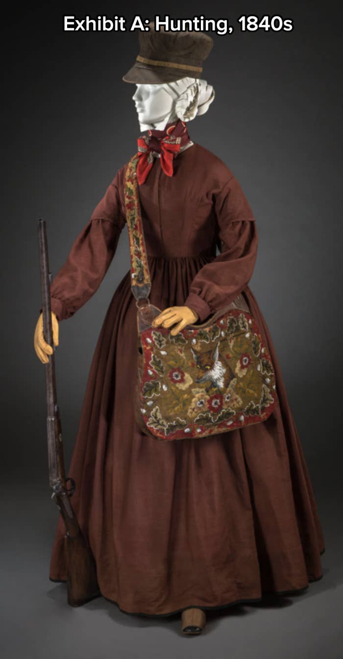 A mannequin is dressed in Victorian-era hunting attire, featuring a brown long-sleeved dress, floral shoulder bag, and holding a rifle
