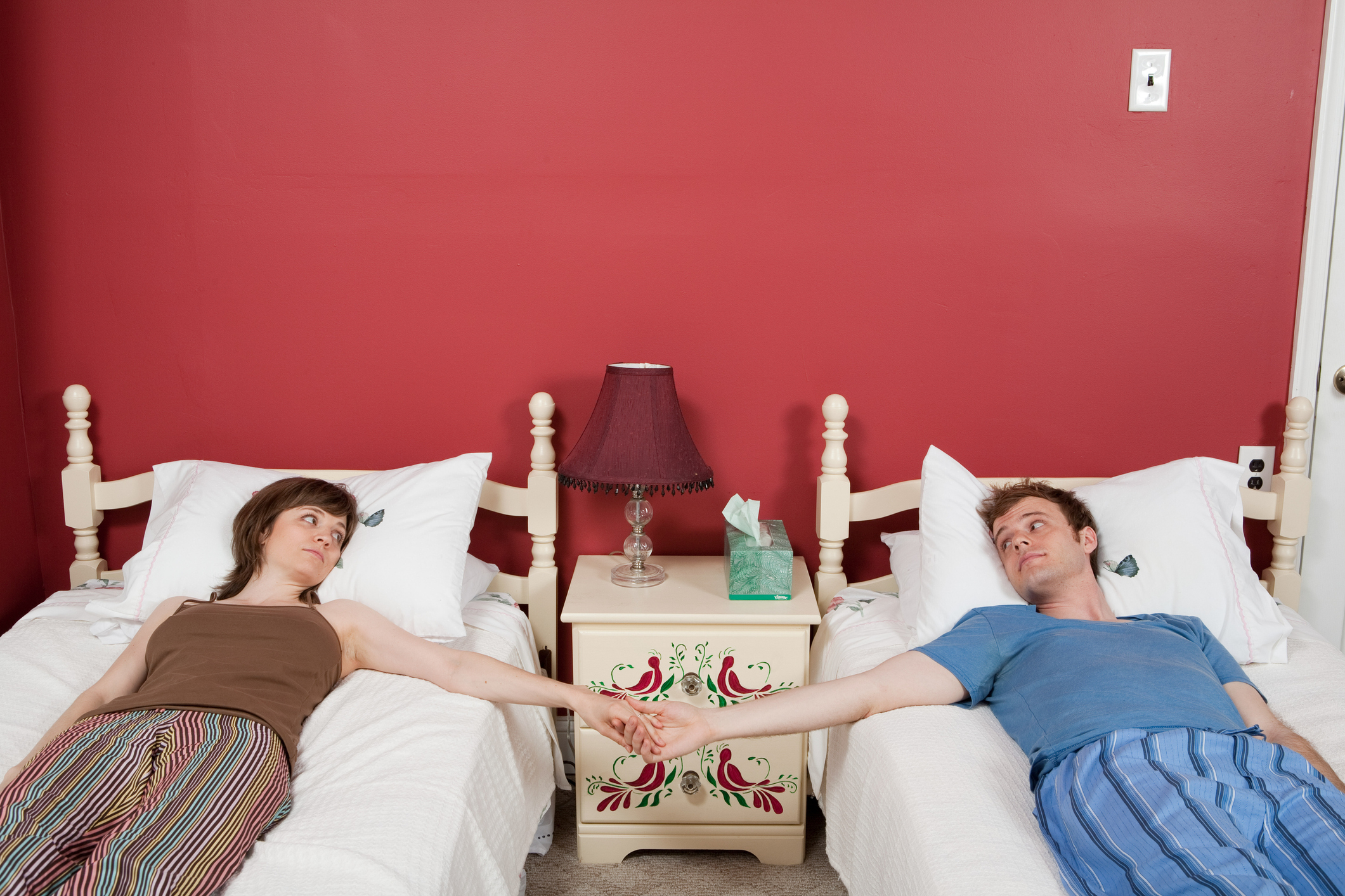 A man and woman lying in separate twin beds while holding hands in the space between beds