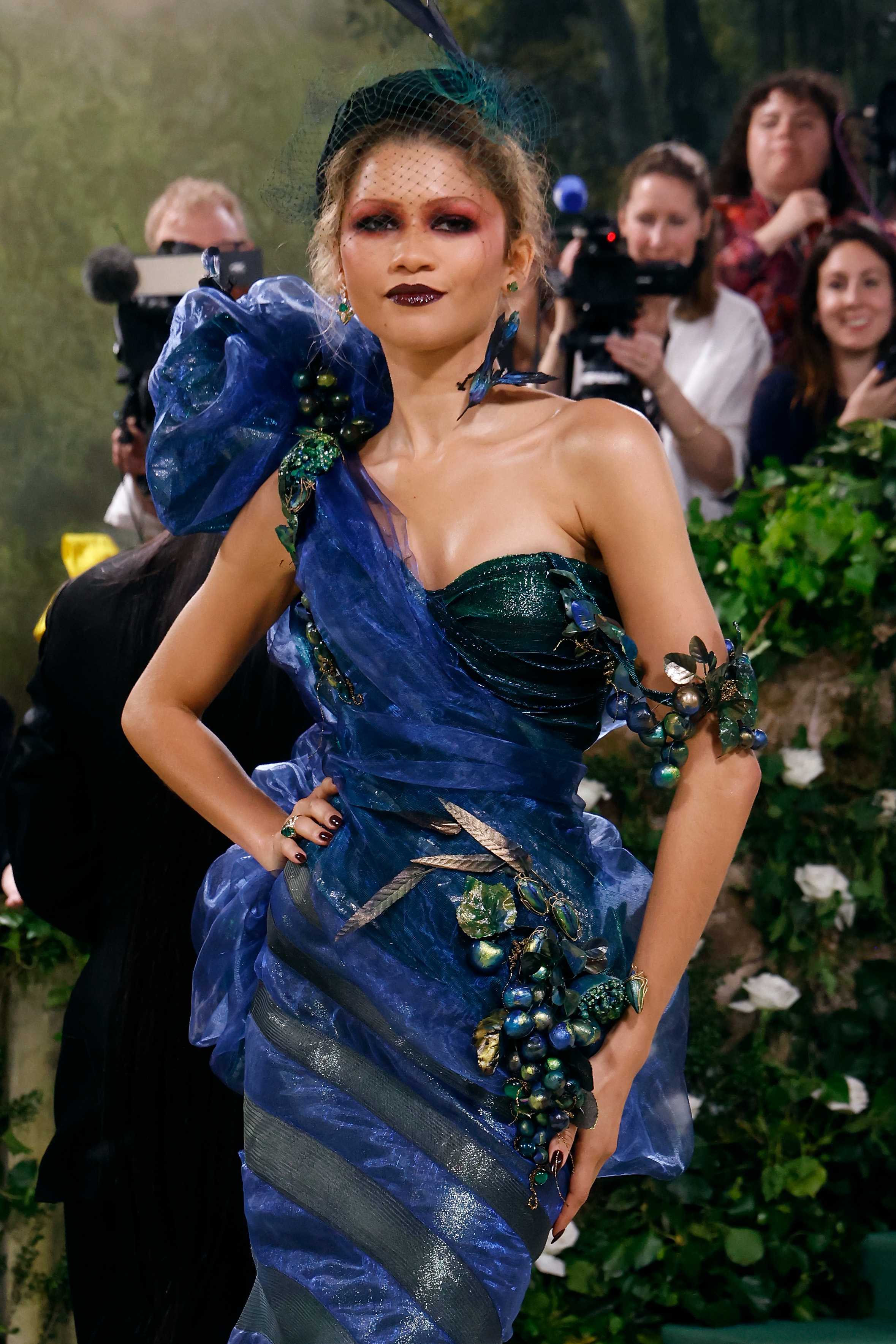 Zendaya in a dramatic, elaborate gown with floral details and accessorized with a headpiece on the red carpet, posing confidently for photographers