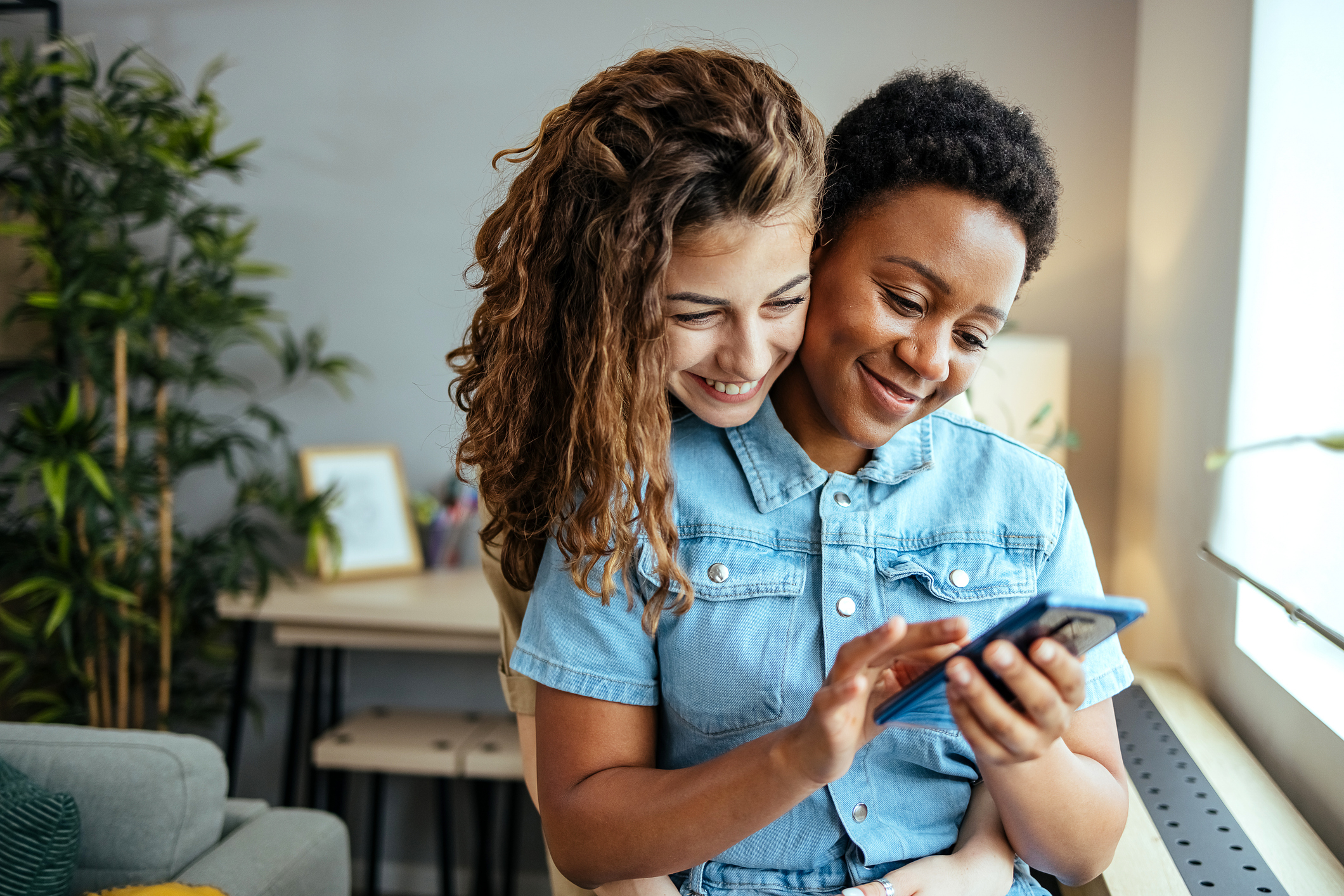 A smiling woman looks over another woman's shoulder while she is typing on her smart phone