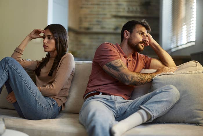 A man and a woman sit on a couch, both looking away from each other with tense expressions, suggesting a disagreement or argument