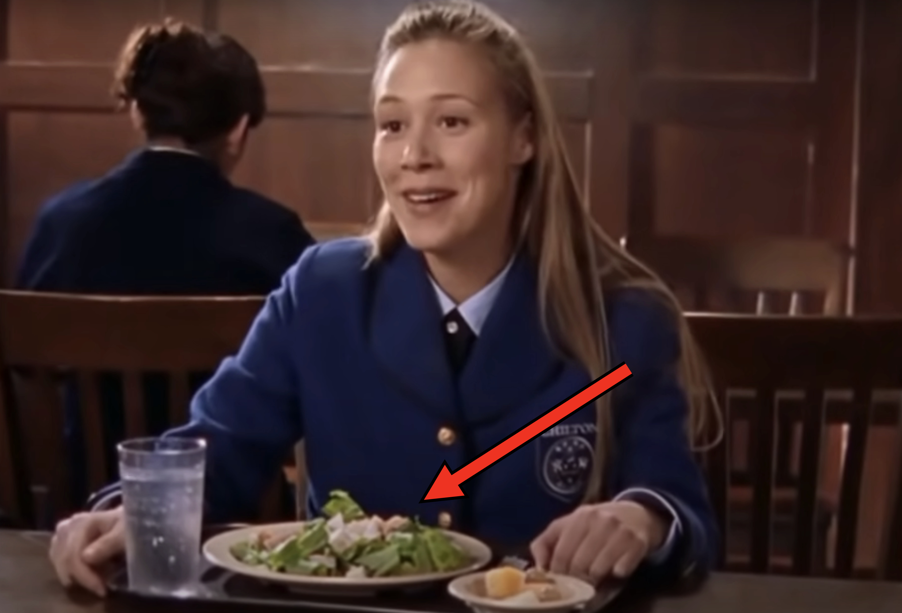 Woman in a school uniform with &quot;CHCETON&quot; badge, smiling while seated at a table with a meal of salad, bread, and a glass of water