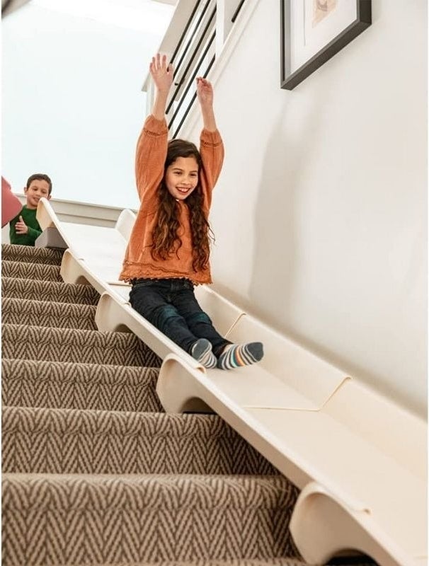 A child slides down a plastic slide on stairs, smiling with arms raised, while another child watches from behind.