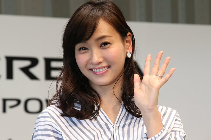 女性が笑顔で手を振っている。彼女は白と黒のストライプのシャツを着用しています。
