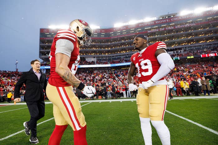 George Kittle and Deebo Samuel of the San Francisco 49ers on the field at Levi's Stadium, with a man in a suit walking in the background