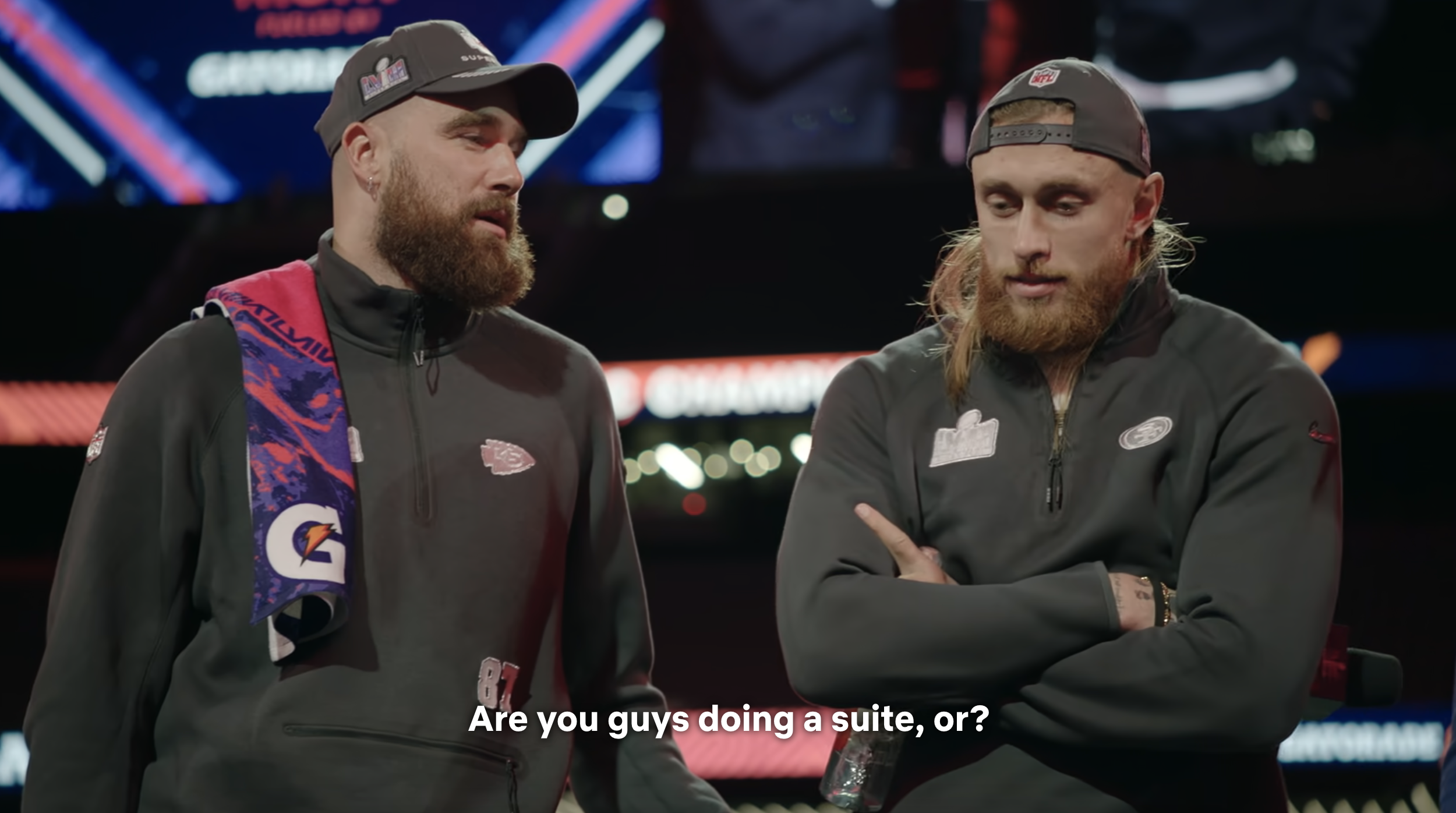 Travis Kelce and George Kittle at a media event, both wearing sports merchandise jackets. Caption: "Are you guys doing a suite, or?"