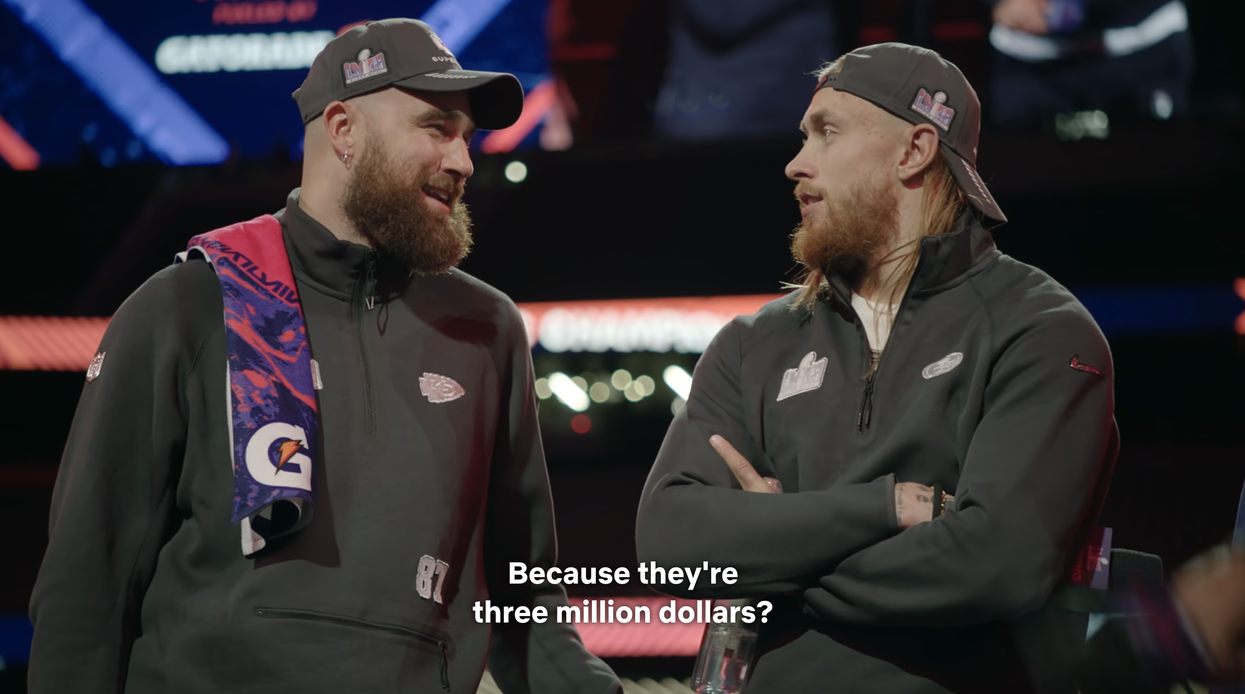 Travis Kelce and George Kittle on stage wearing team jackets and caps, engaged in conversation, captured at a media event. Subtitles read: "Because they're three million dollars?"