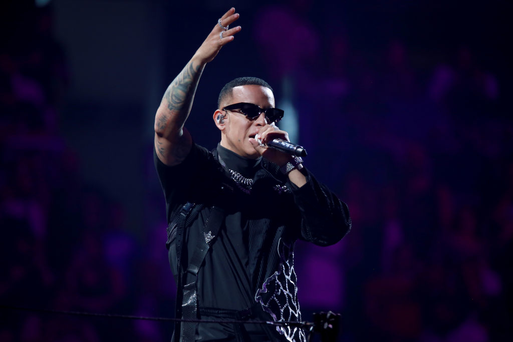 Daddy Yankee performs on stage, wearing sunglasses and a stylish outfit with intricate designs. He has one arm raised, holding a microphone
