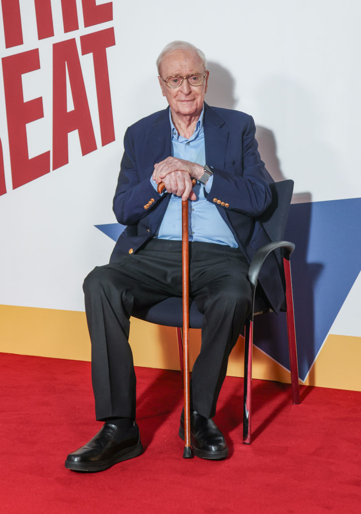Michael Caine sits on a chair with a cane, wearing a suit jacket and dress pants, on a red carpet backdrop