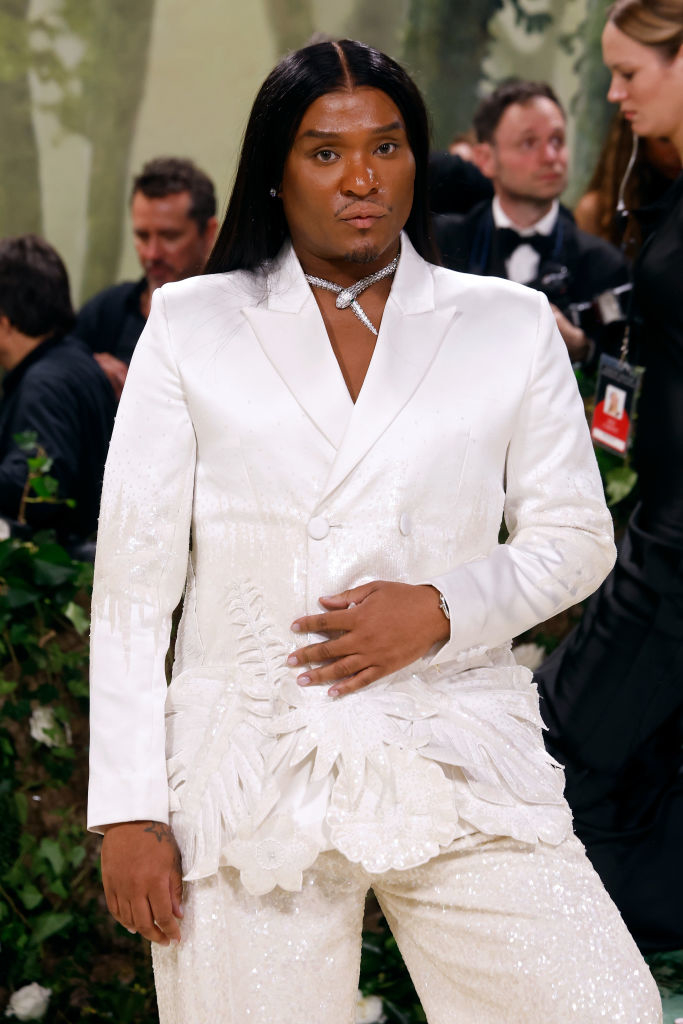 Law in an embroidered suit with leaf patterns and a diamond necklace on a red carpet. Background includes several people and greenery